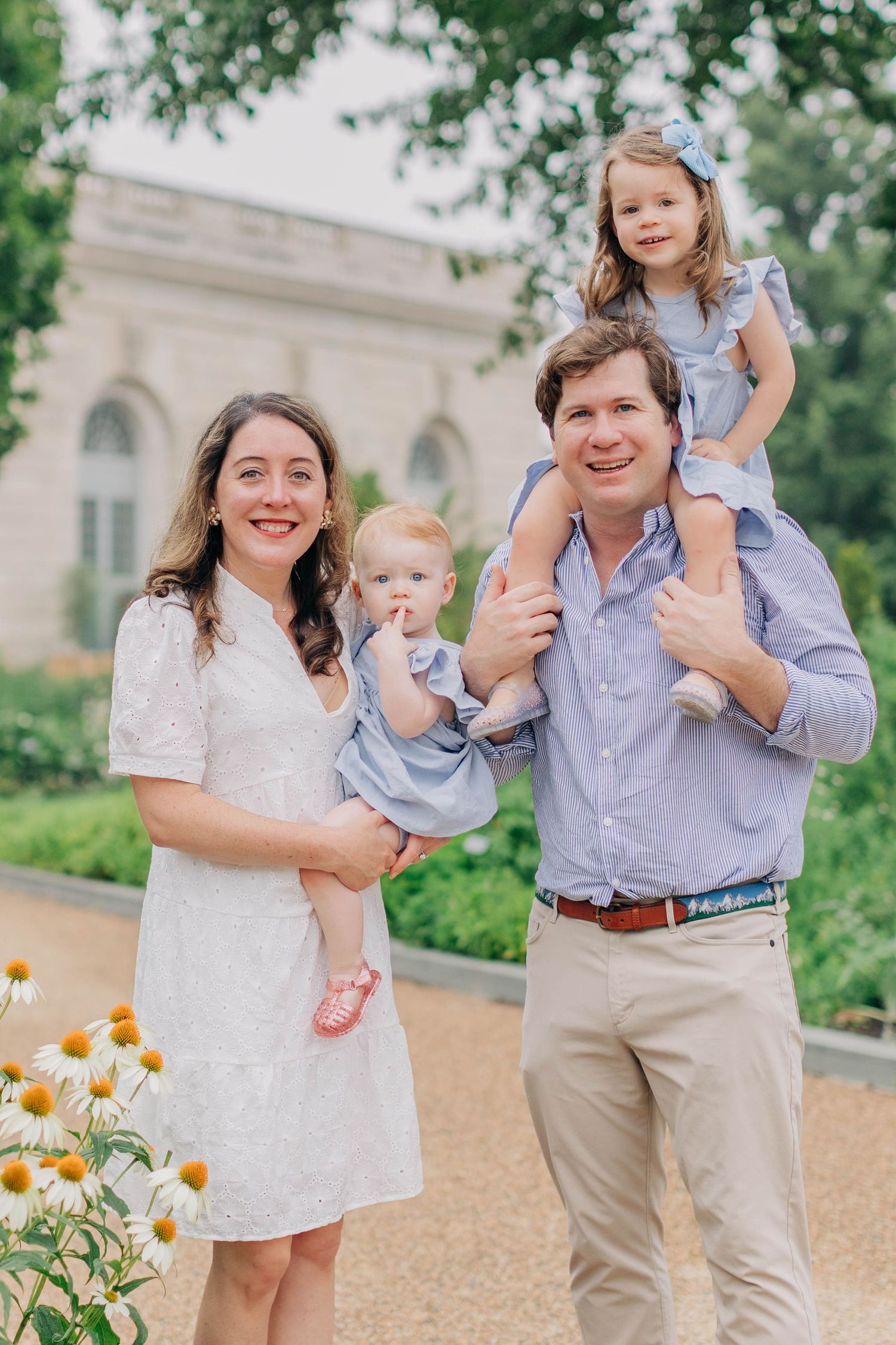 Happy family of four posing outdoors in a garden, parents holding two young daughters in matching blue dresses.