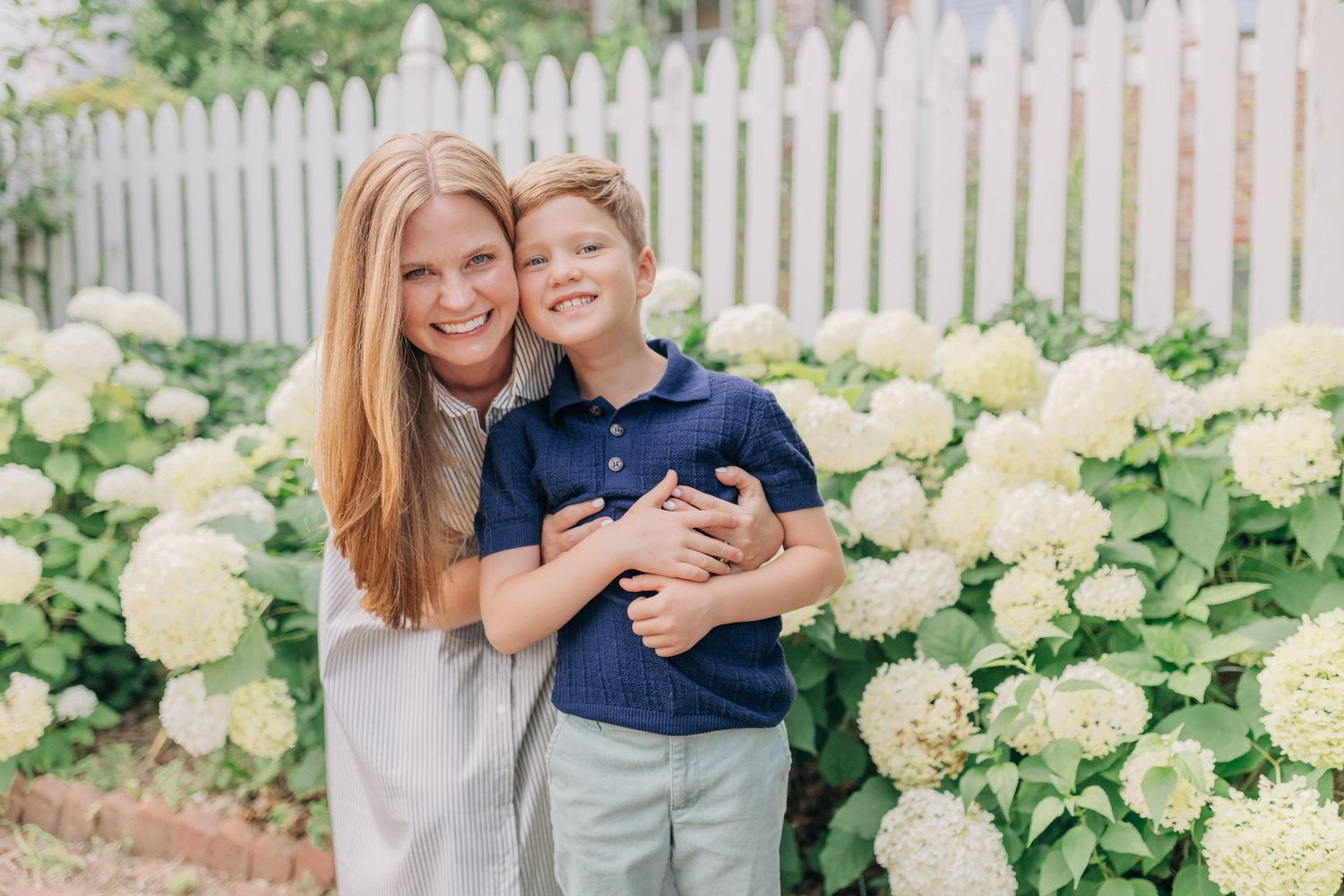 A smiling woman hugs a young boy among white hydrangea bushes near a white picket fence outdoors.