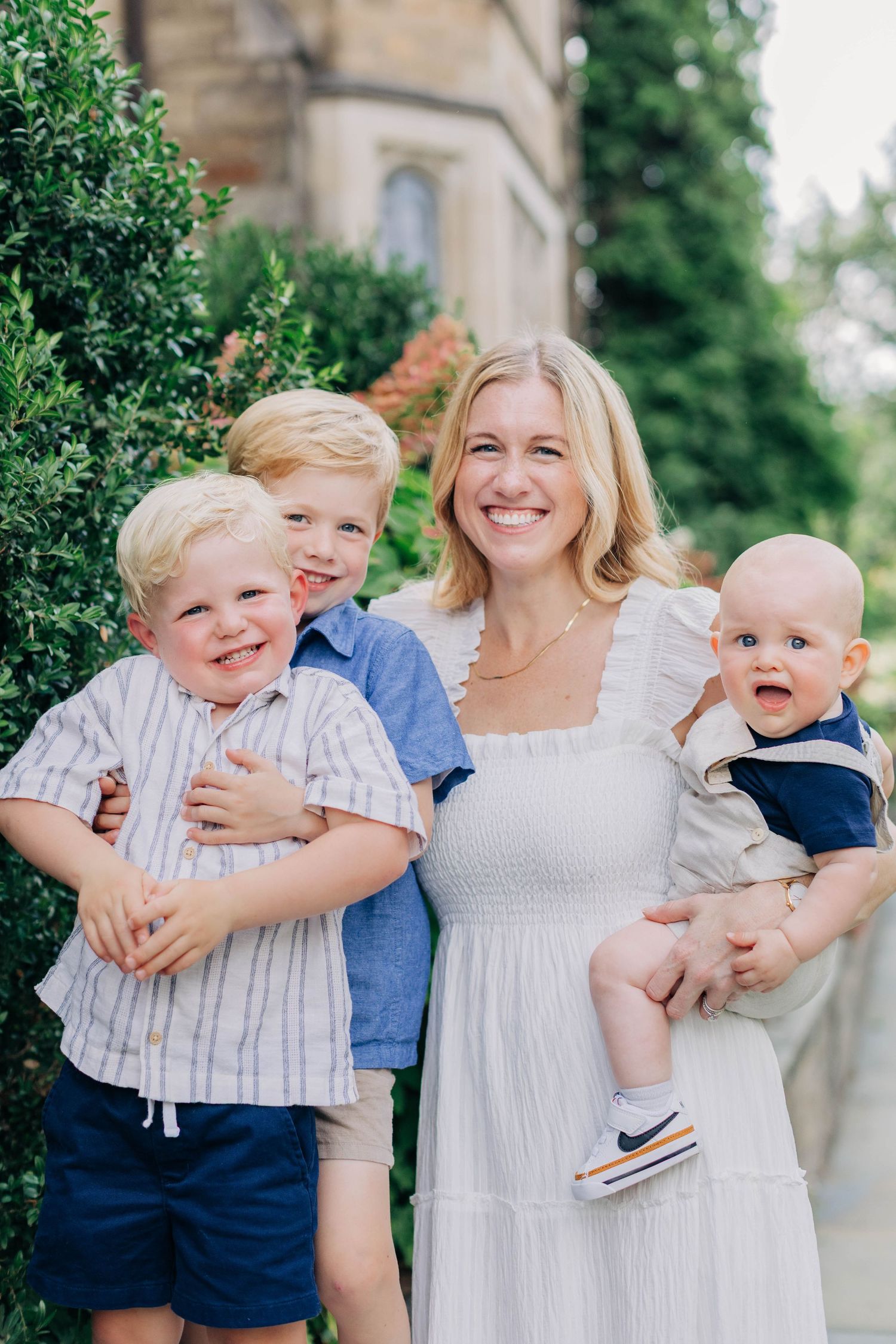 Smiling blonde mother in white dress holding three young boys outdoors near green hedges.