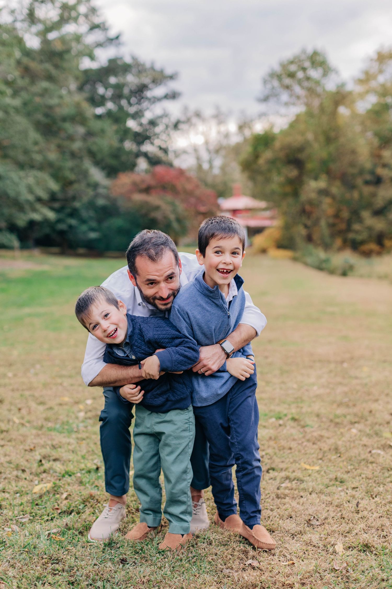 A father hugs his two young sons in a lush green park during autumn, all smiling joyfully.