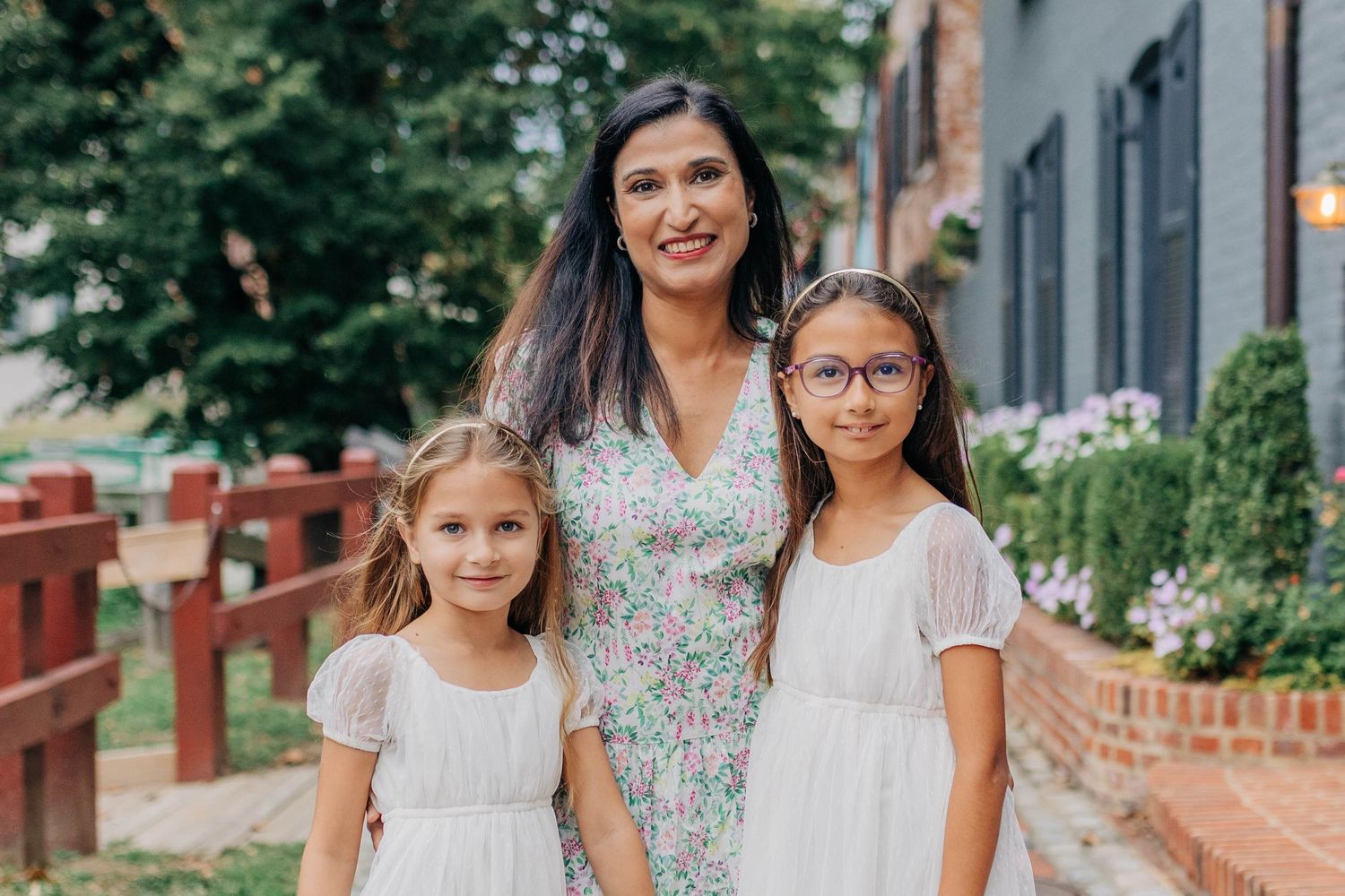 A woman in a floral dress poses outdoors with two young girls in white dresses, smiling together near a brick wall.