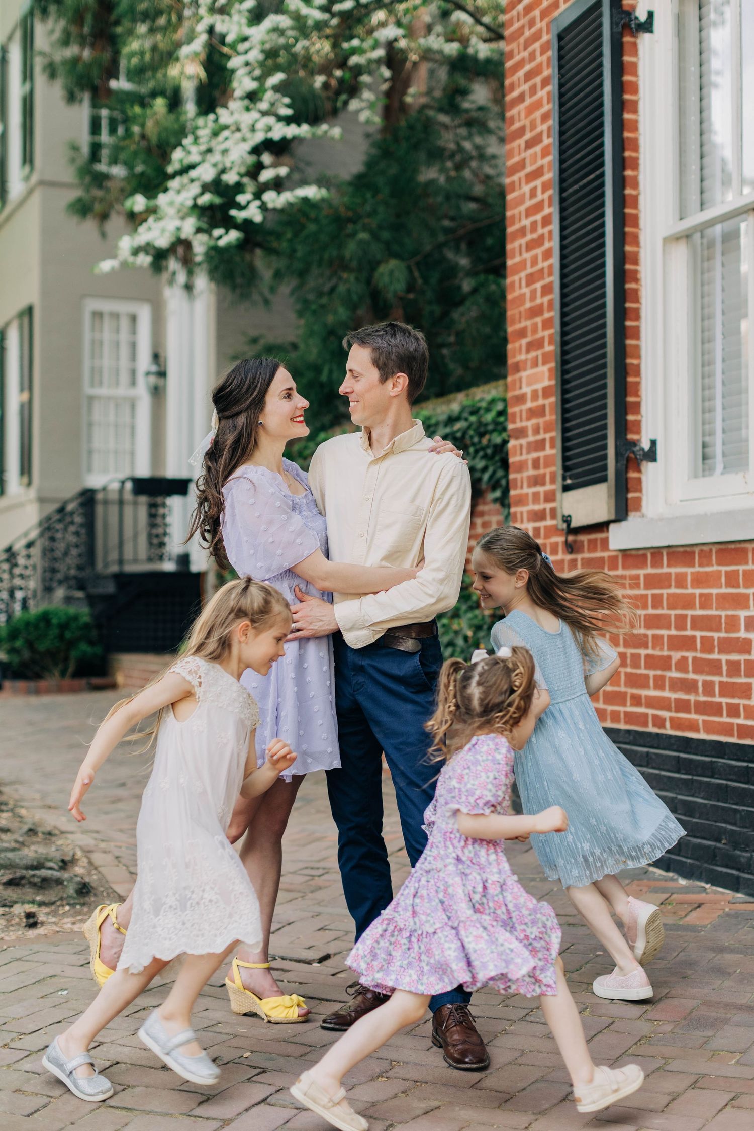 A happy family with three young girls in dresses dancing around their smiling parents outside a brick home.