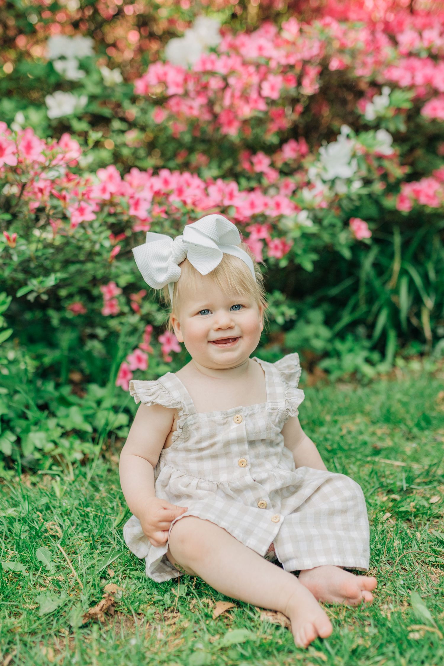 A baby in a linen dress and white bow sits on grass surrounded by pink azalea bushes.