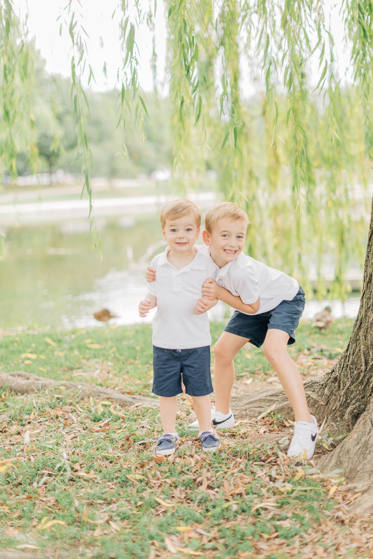 Two young children playing and running together under a weeping willow tree near a lake.