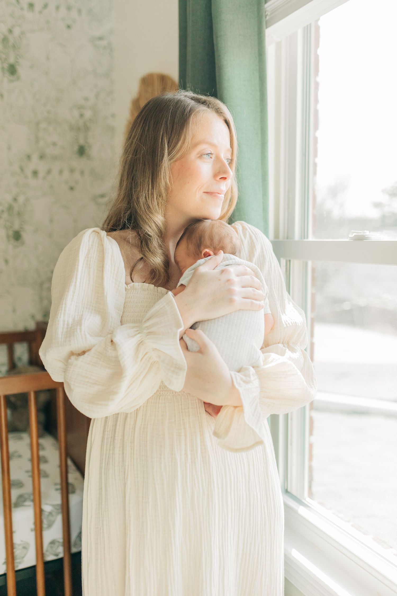 Sunlit nursery scene with green curtains and wooden crib beside bright window.