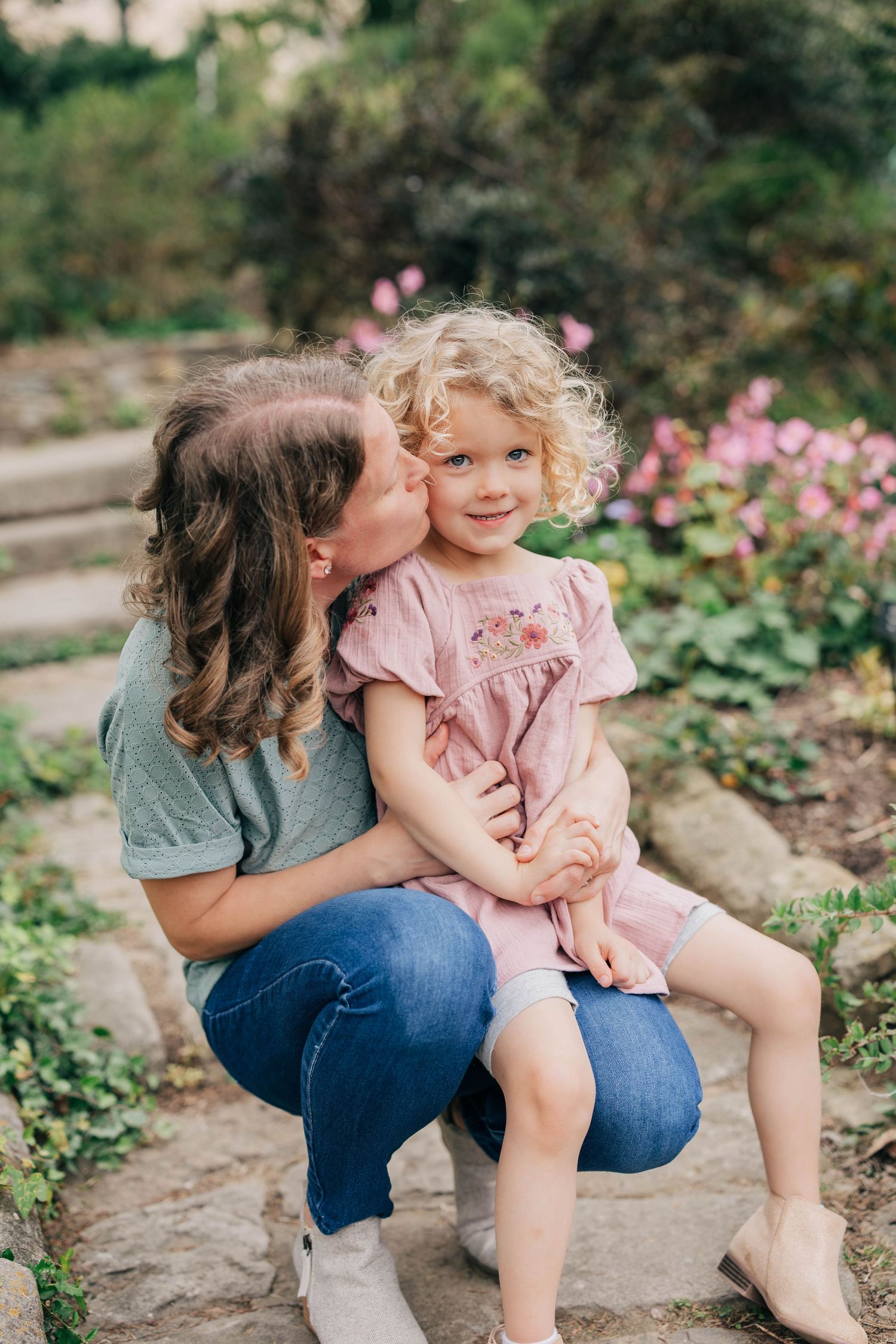 A mother in a gray tee and jeans kisses the cheek of her smiling curly-haired daughter on stone steps near pink flowers.