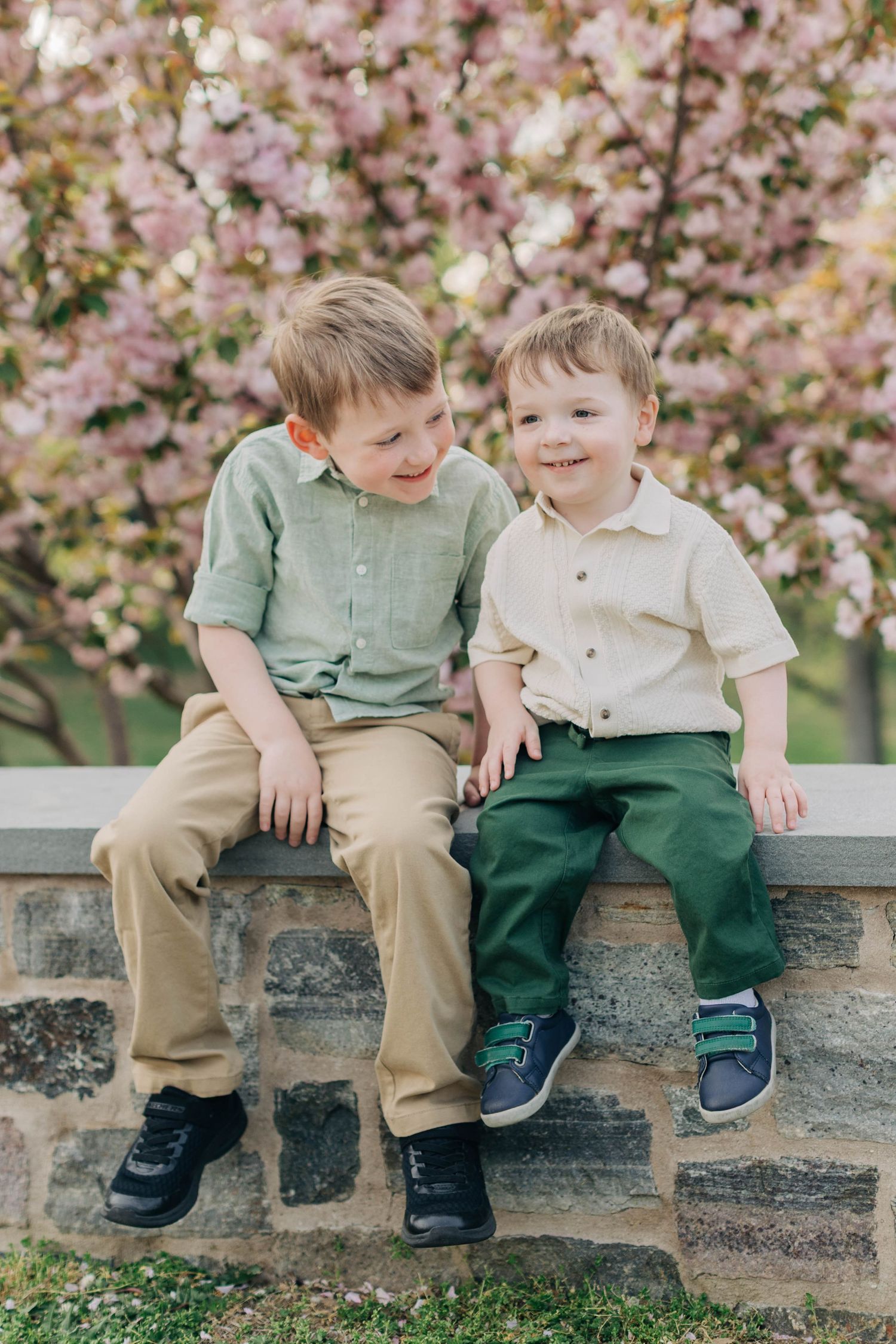Two young brothers sitting together on a stone wall in front of pink cherry blossom trees in spring.