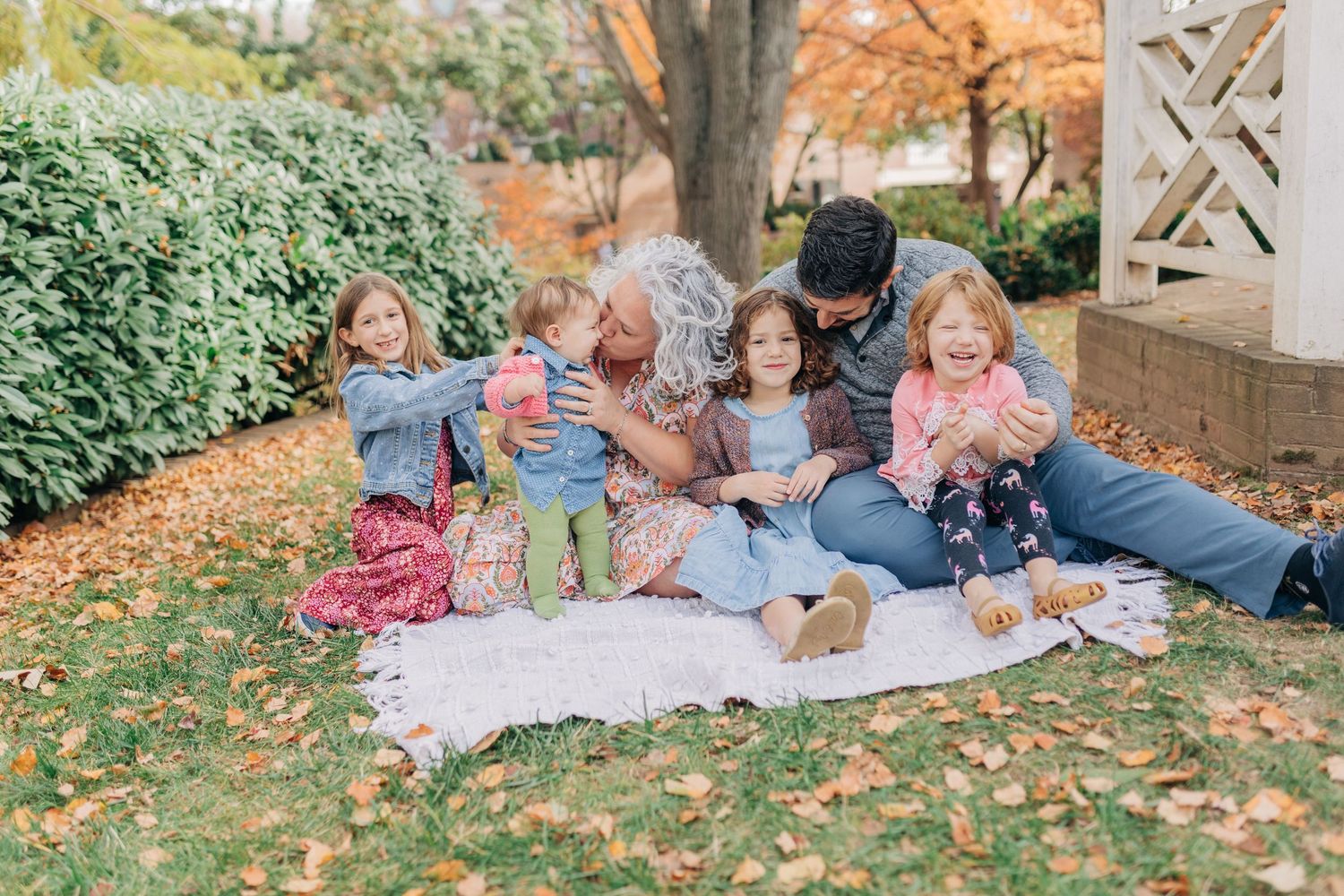 Family sitting on a blanket outdoors in autumn, laughing and enjoying time together under a tree with fall foliage.