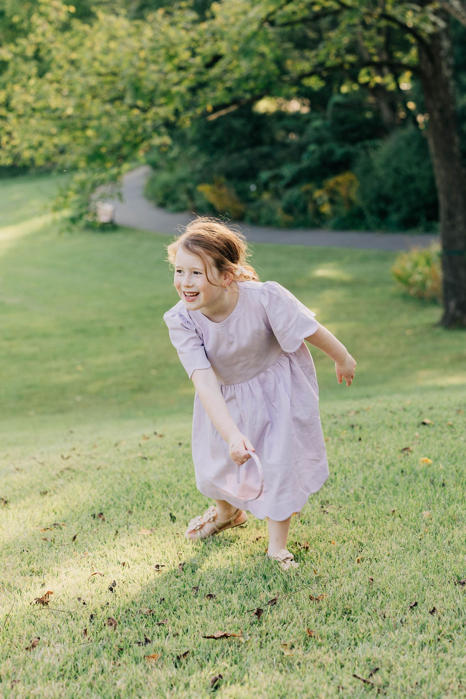 A joyful young girl in a lavender dress runs and plays on a lush green lawn in a sunny garden.
