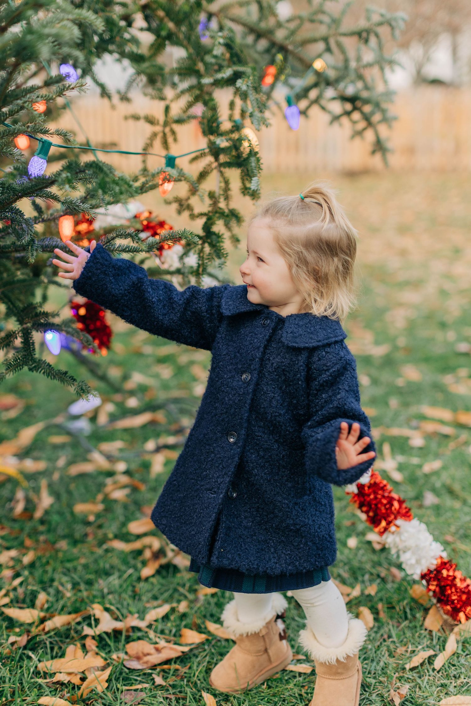 Toddler girl in navy coat decorating outdoor Christmas tree with red ornaments and colorful lights in autumn yard.