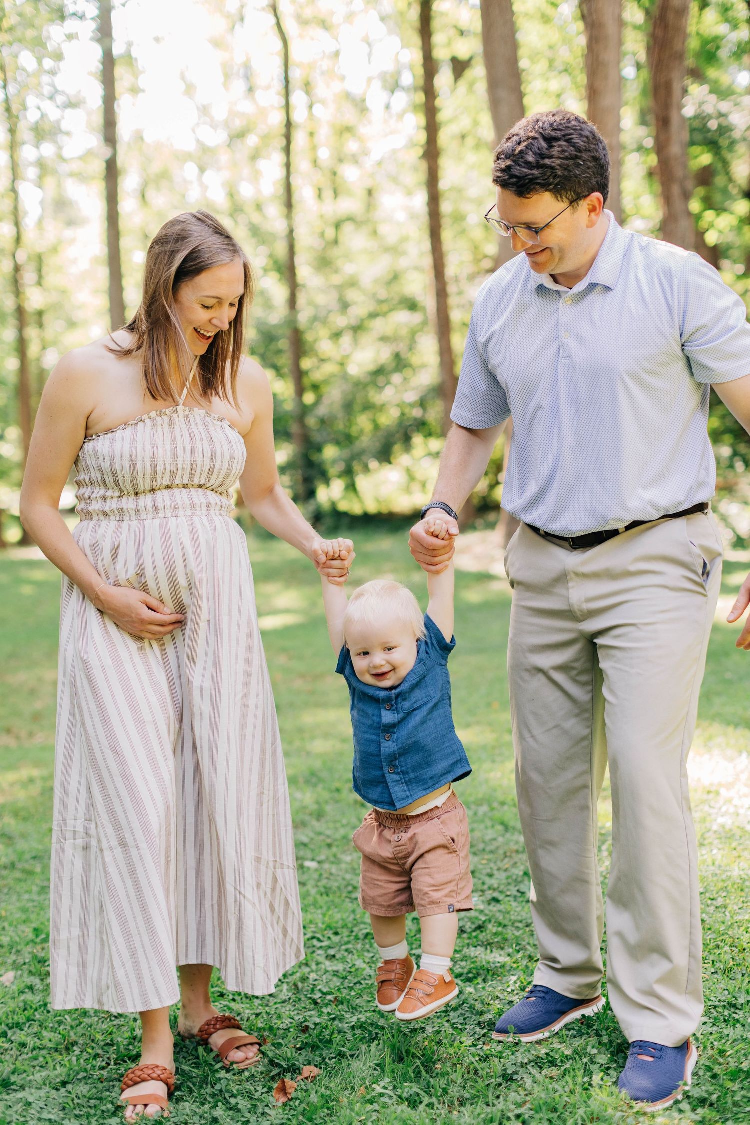 Pregnant woman and man hold toddler boy's hands in sunny green park, family maternity photo session outdoors.