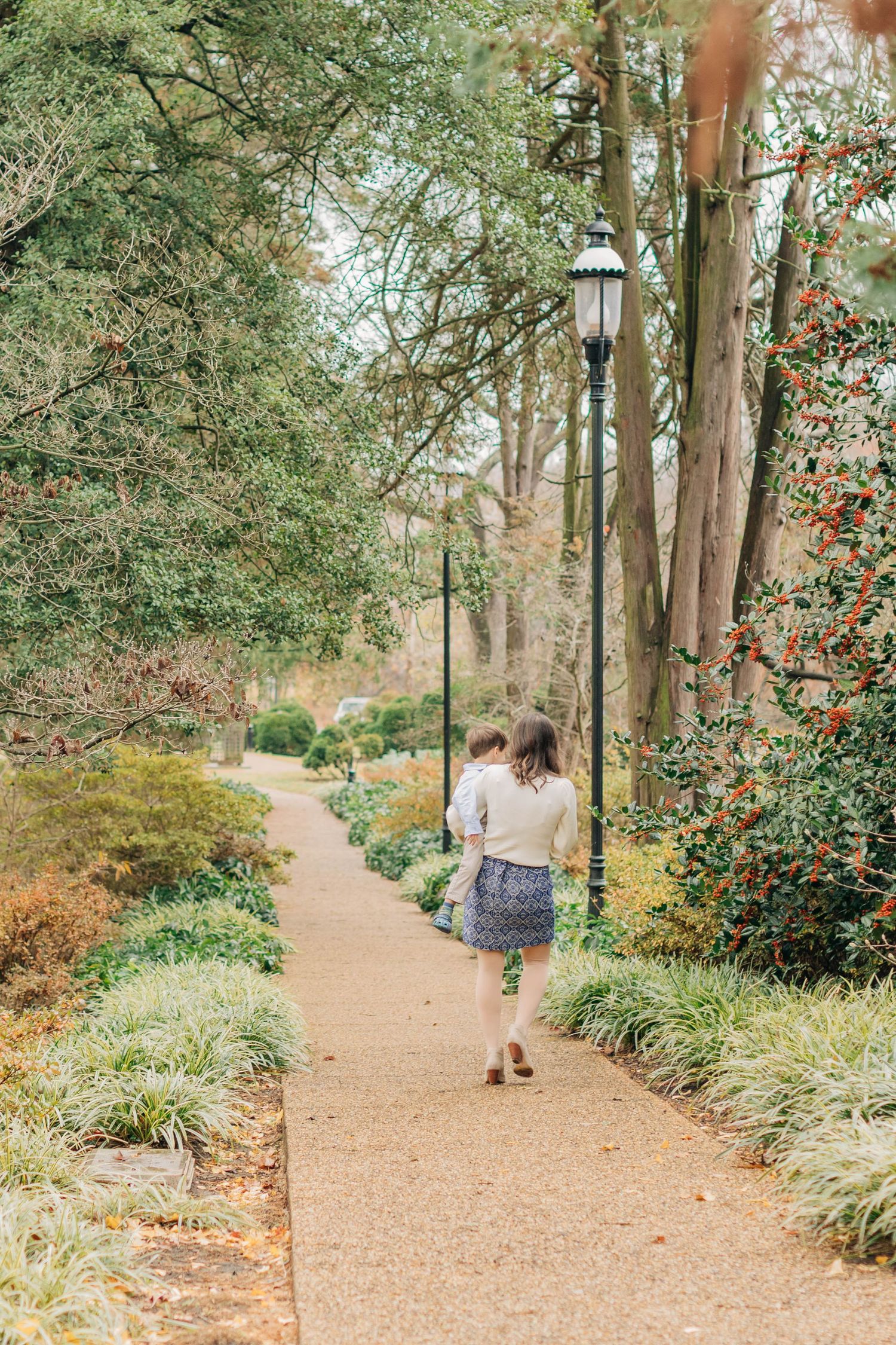 A person walks along a scenic garden path lined with lampposts and autumn foliage.
