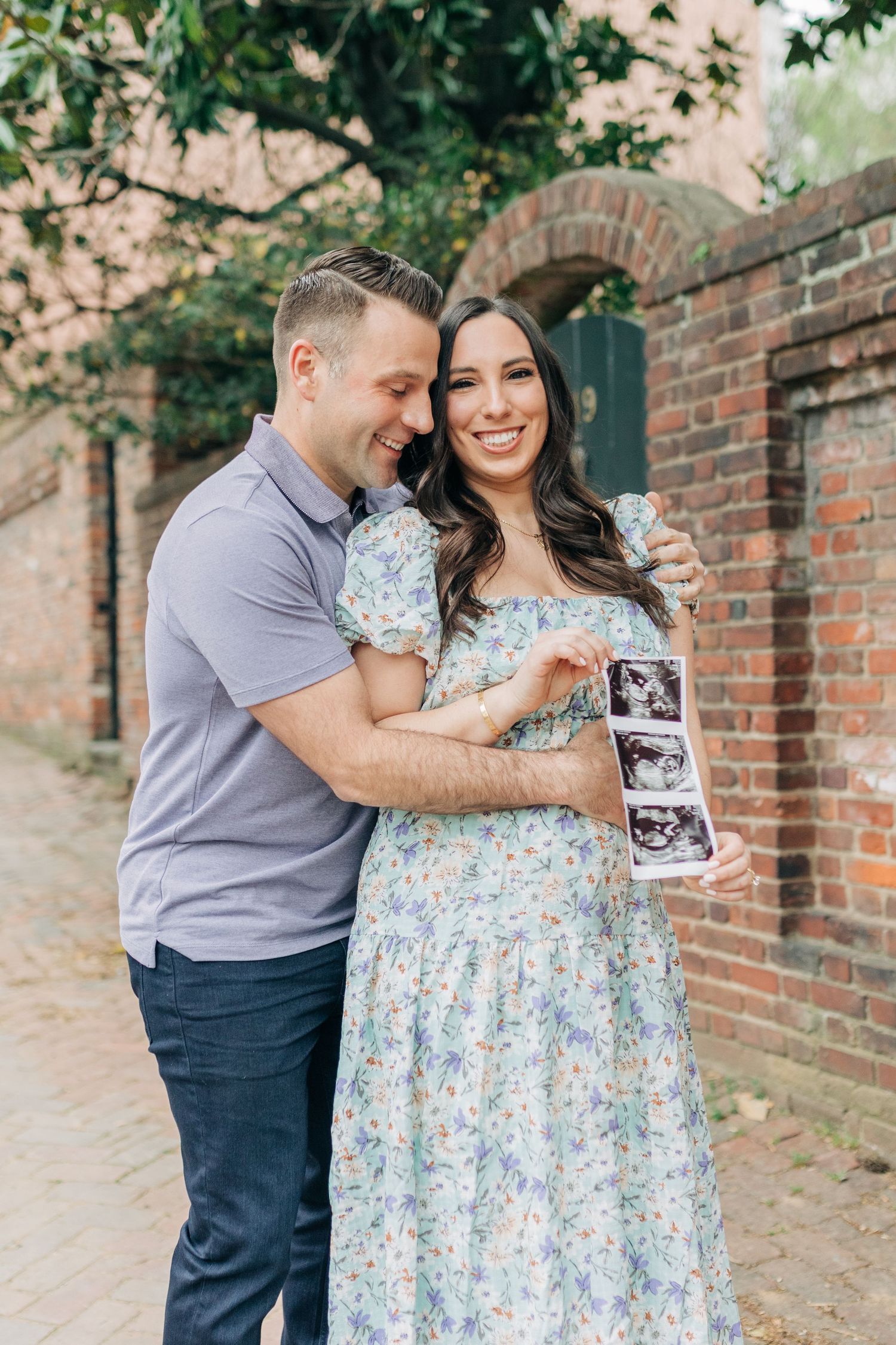 A couple shares happy moments while holding ultrasound photos on a historic brick pathway.