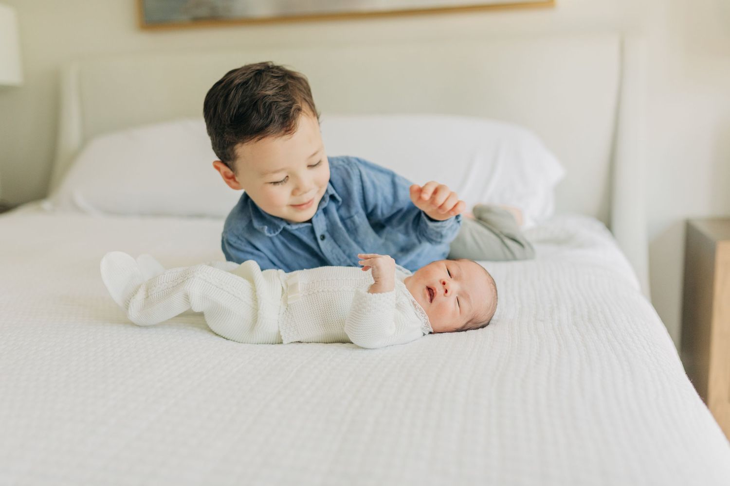 Two siblings share sweet interactions on a white bed in a minimalist bedroom.