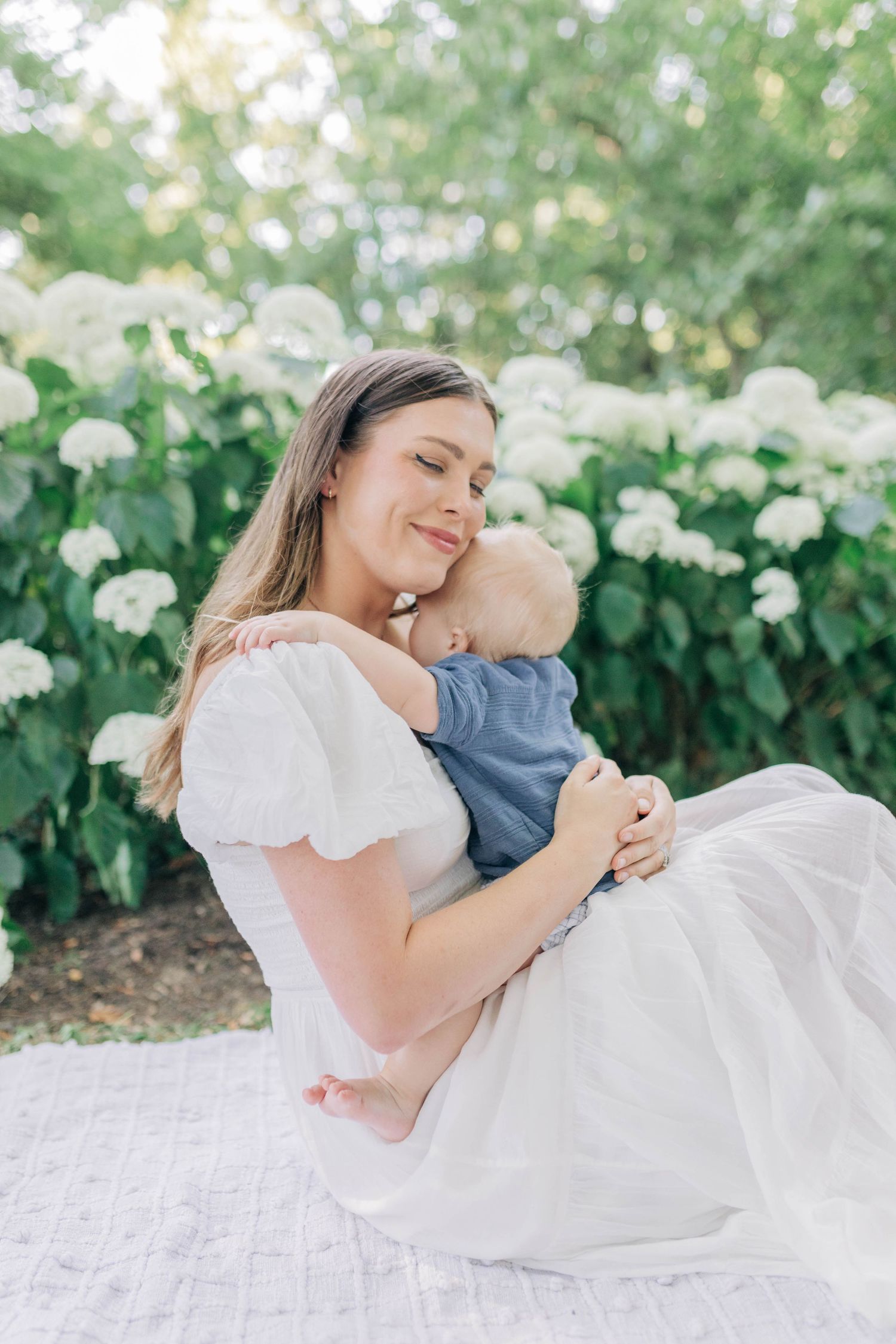 Mother and baby share loving embrace on white blanket surrounded by white flowers.