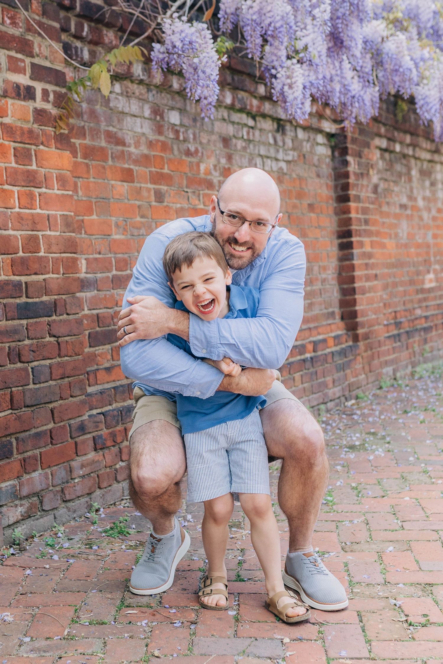 Bald father hugging laughing son in front of brick wall with purple wisteria flowers blooming overhead.