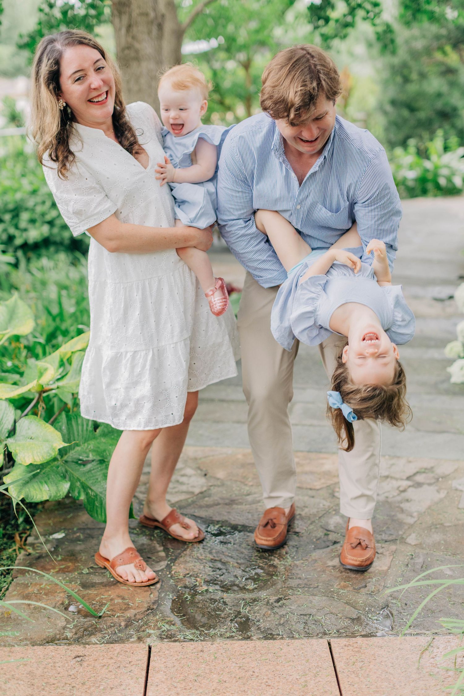 Joyful family of four playing outdoors, father swinging laughing daughter upside down in lush garden.