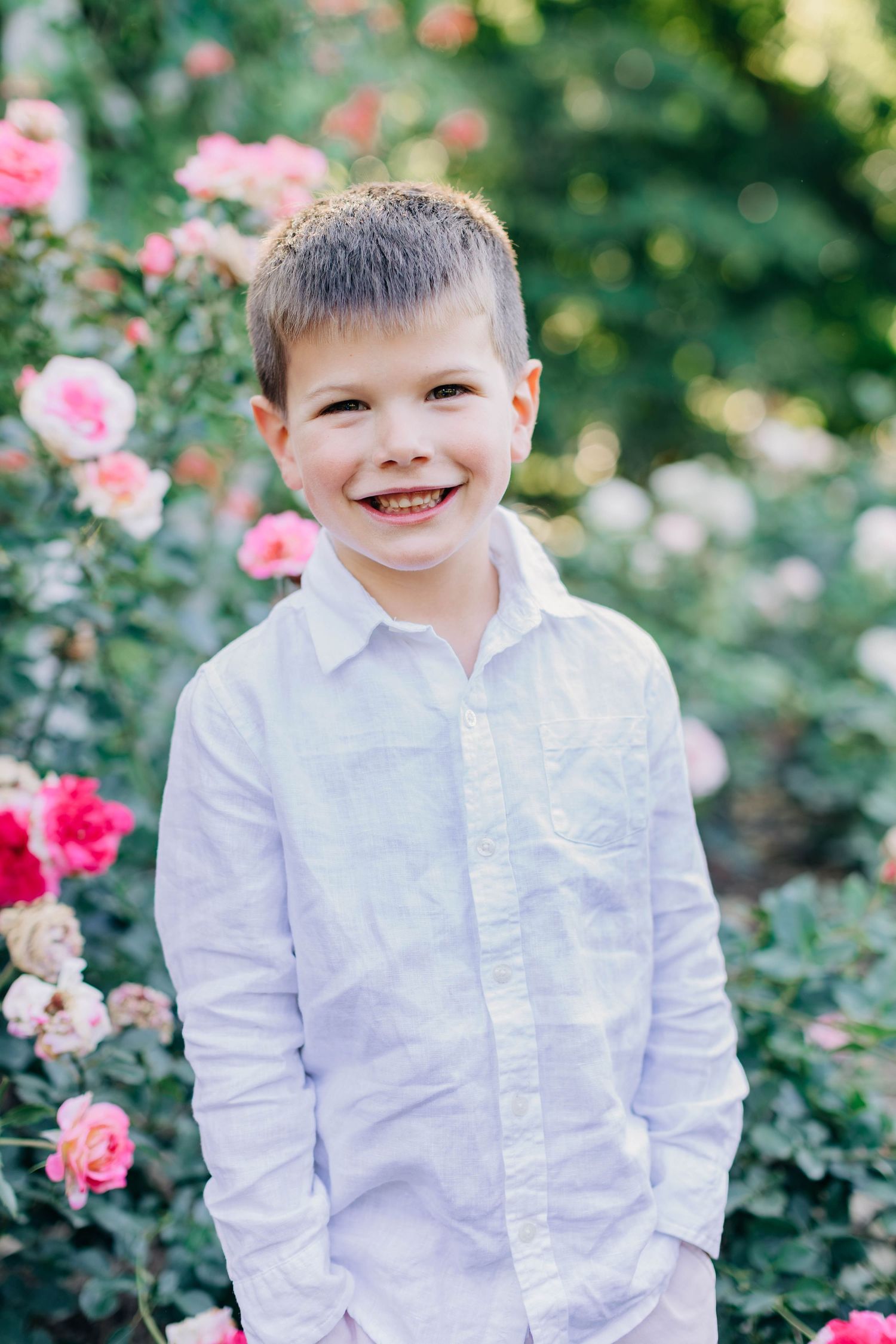 Smiling young boy in white shirt posing in front of pink roses in a garden.