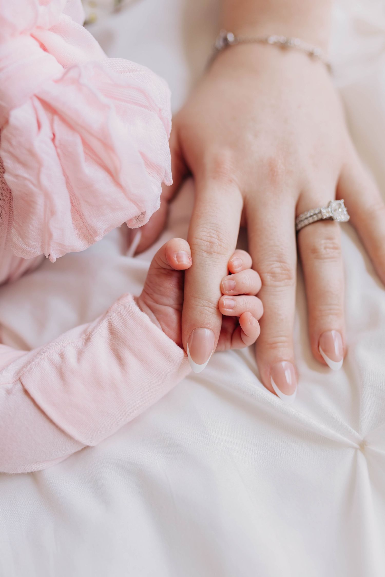 A baby's tiny hand gently gripping a mother's finger, wearing a diamond ring and bracelet, on white bedding.