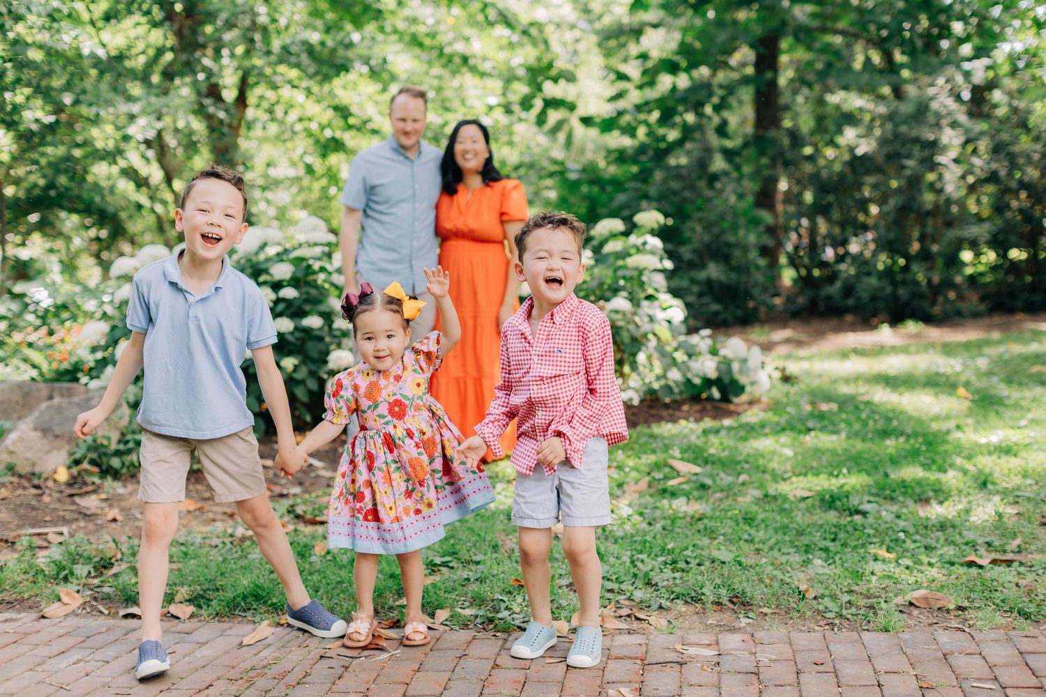 Happy family with three laughing children poses outdoors on brick path surrounded by lush green garden trees.