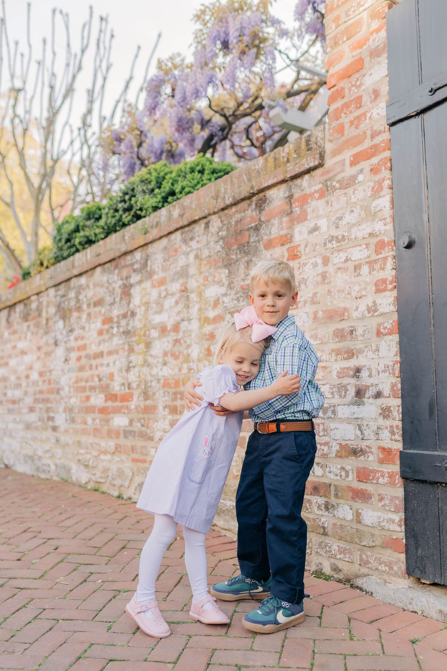 Two young siblings hug beside a brick wall with purple wisteria blooms, dressed in pastel spring outfits.