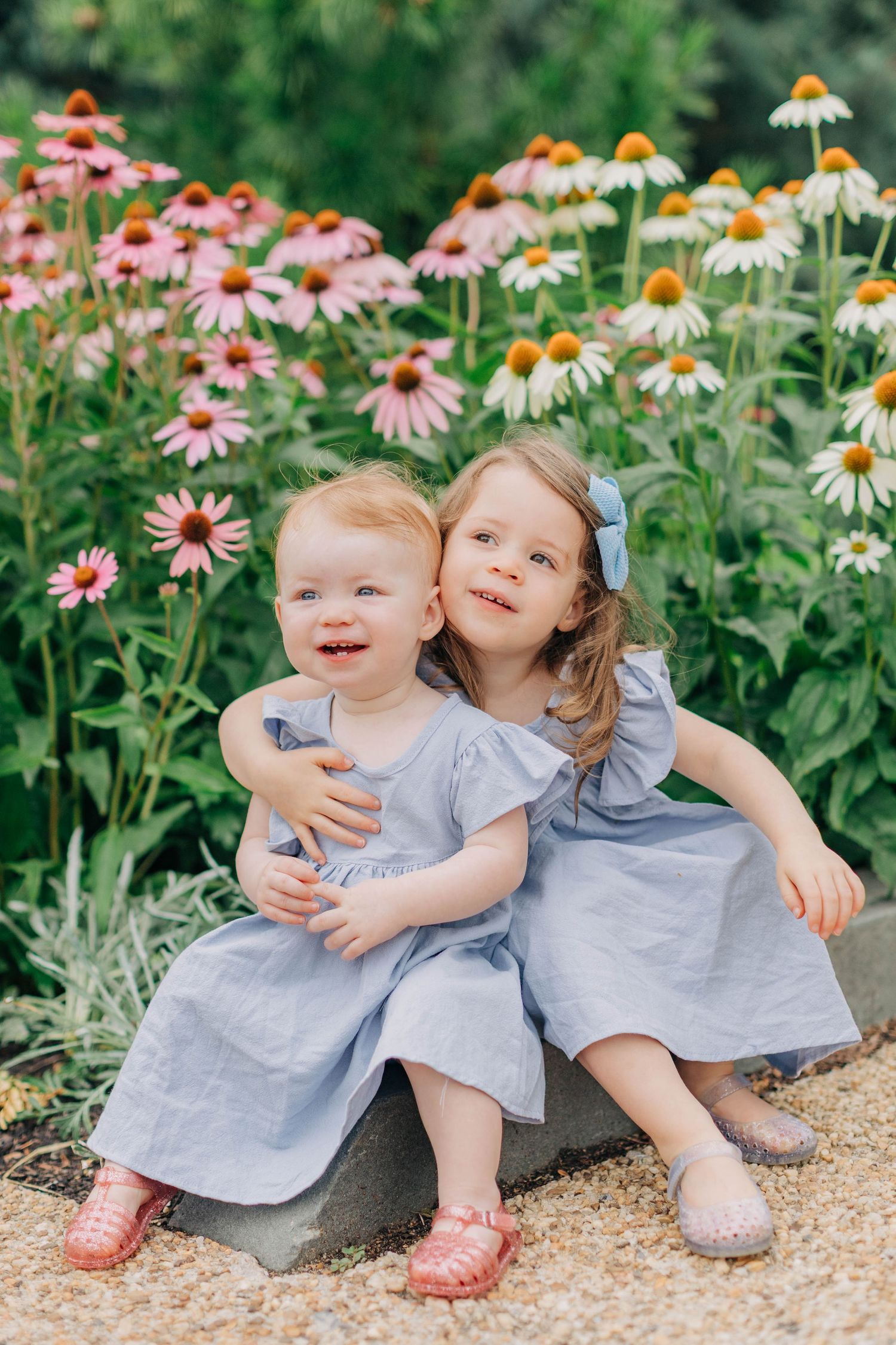 Two young sisters in matching blue dresses embrace in a garden surrounded by blooming pink coneflowers.