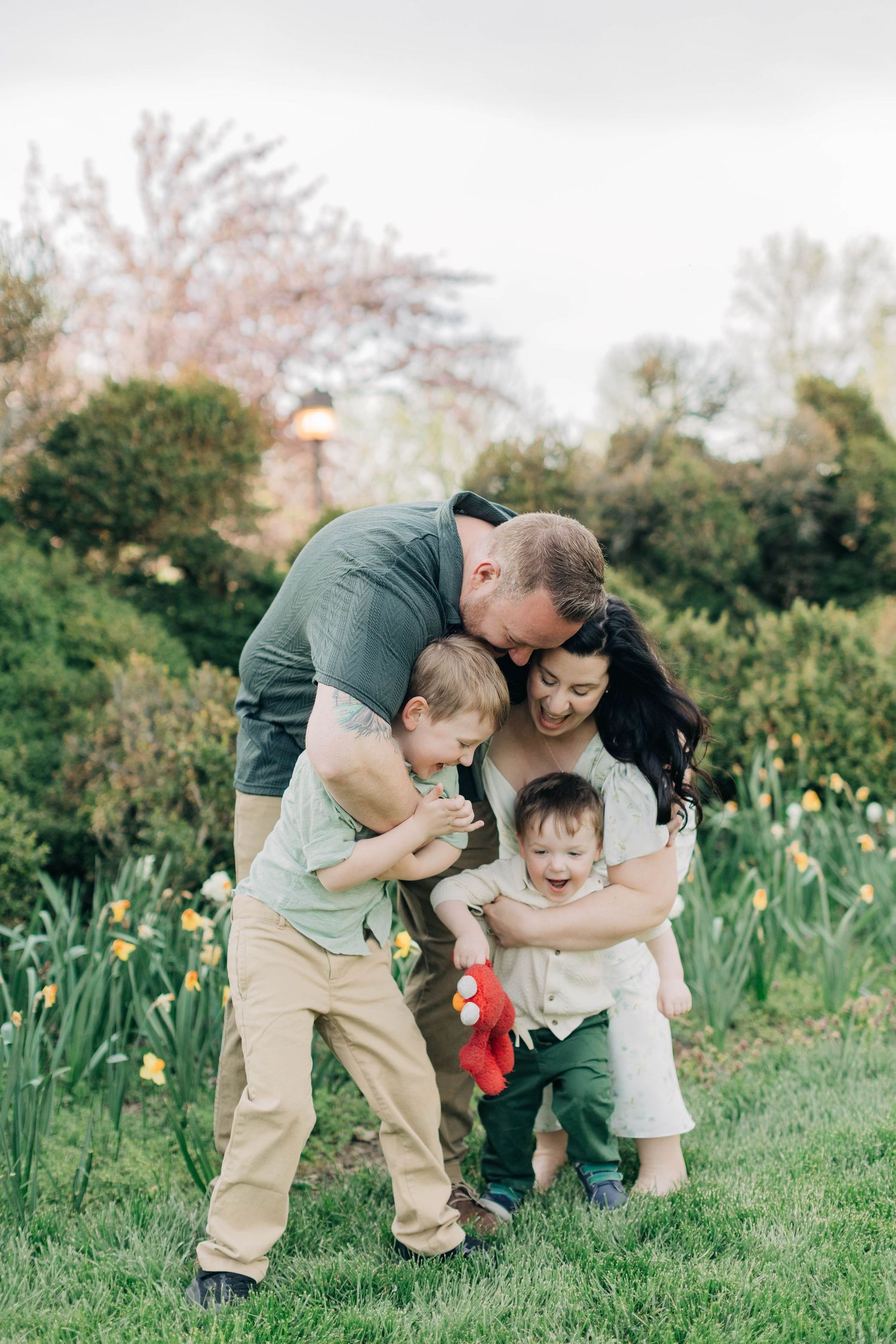 Happy family of four sharing a joyful embrace in a garden with daffodils and cherry blossoms.