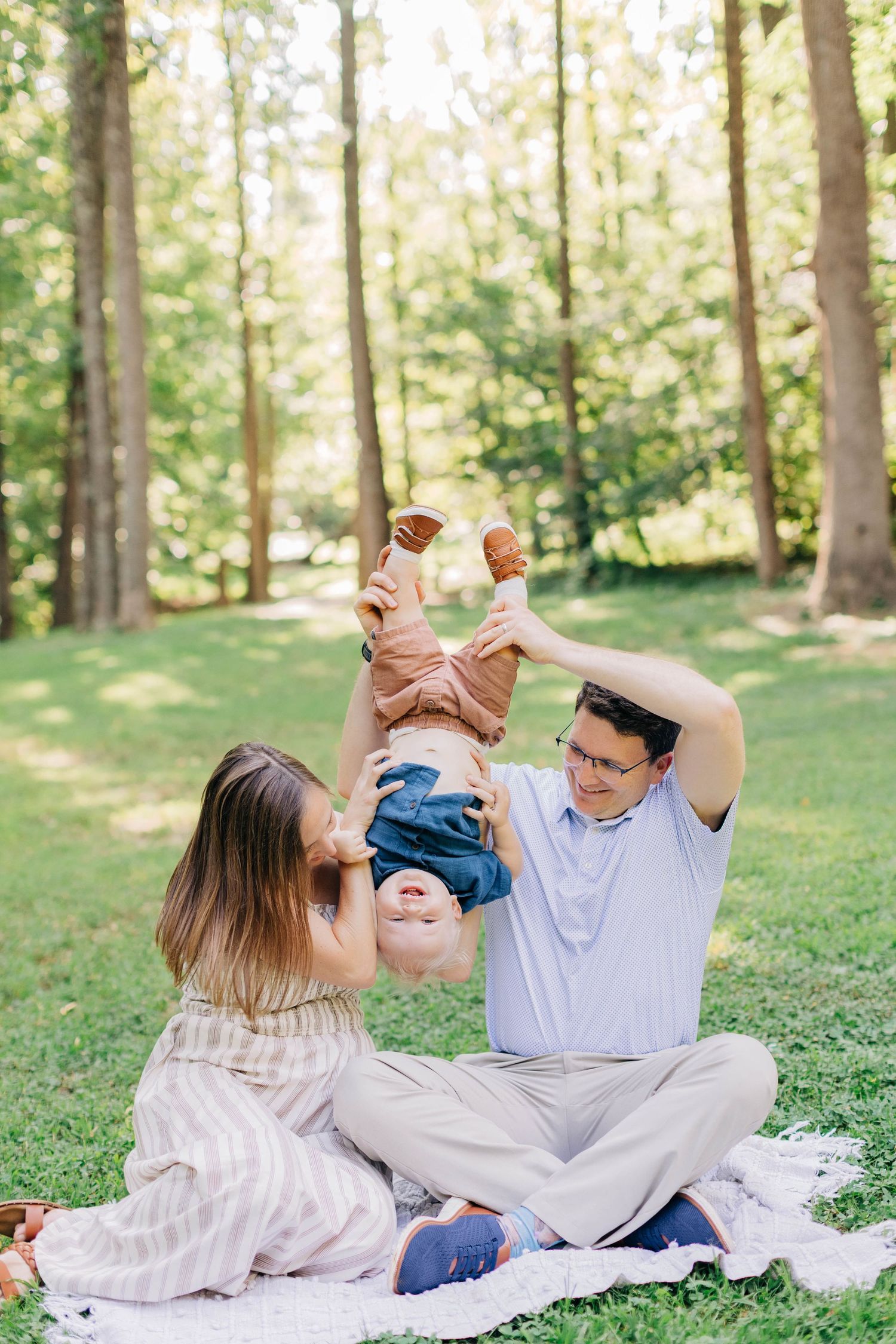 Family enjoying a sunny park picnic, parents playfully lifting their baby upside down on a blanket surrounded by tall trees.