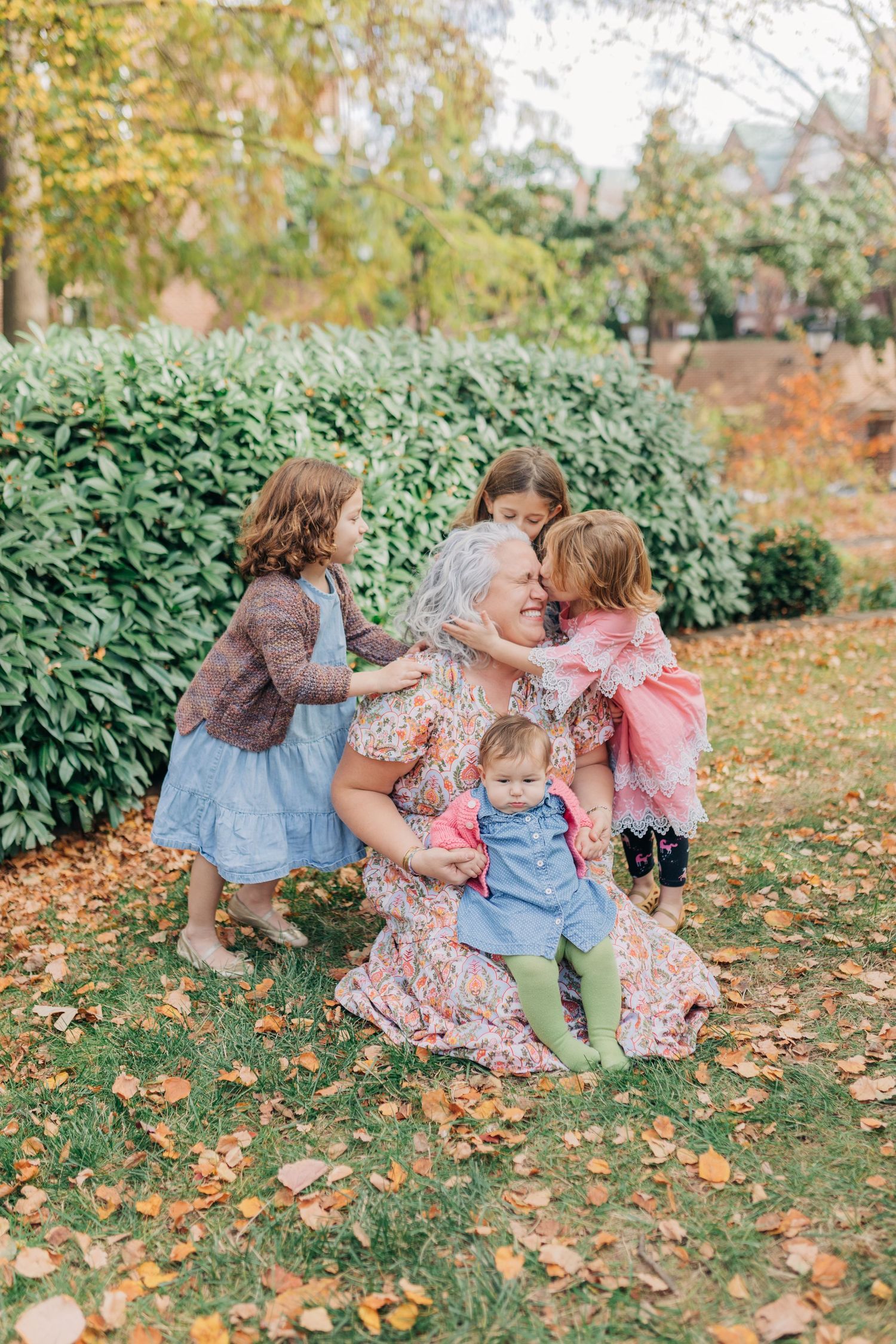 Grandmother kneeling on autumn grass hugging four young granddaughters in colorful dresses outdoors.