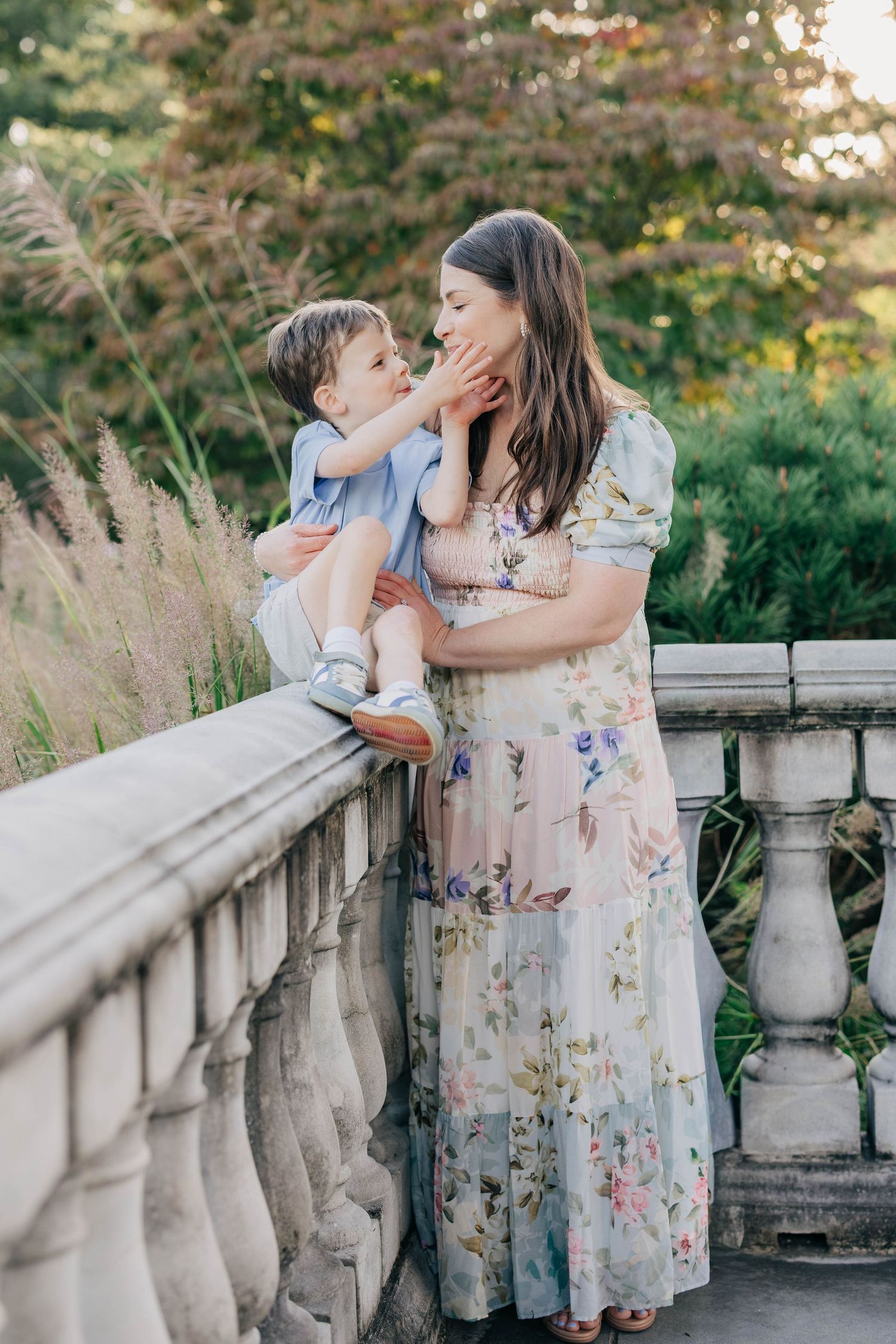 A mother in a floral dress tenderly holds her young son on a stone balustrade.