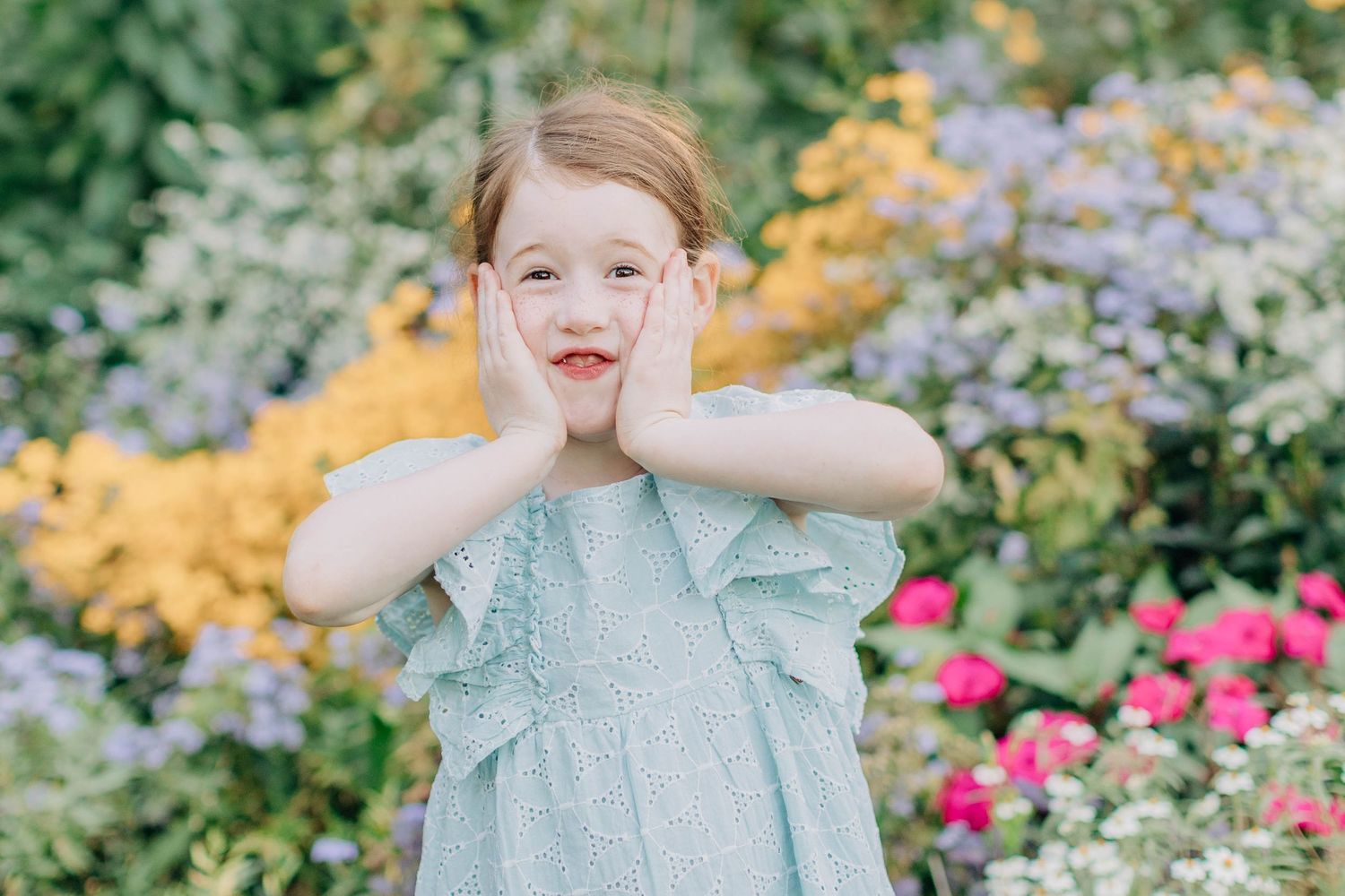 A child in a mint green dress poses playfully in a blooming garden.