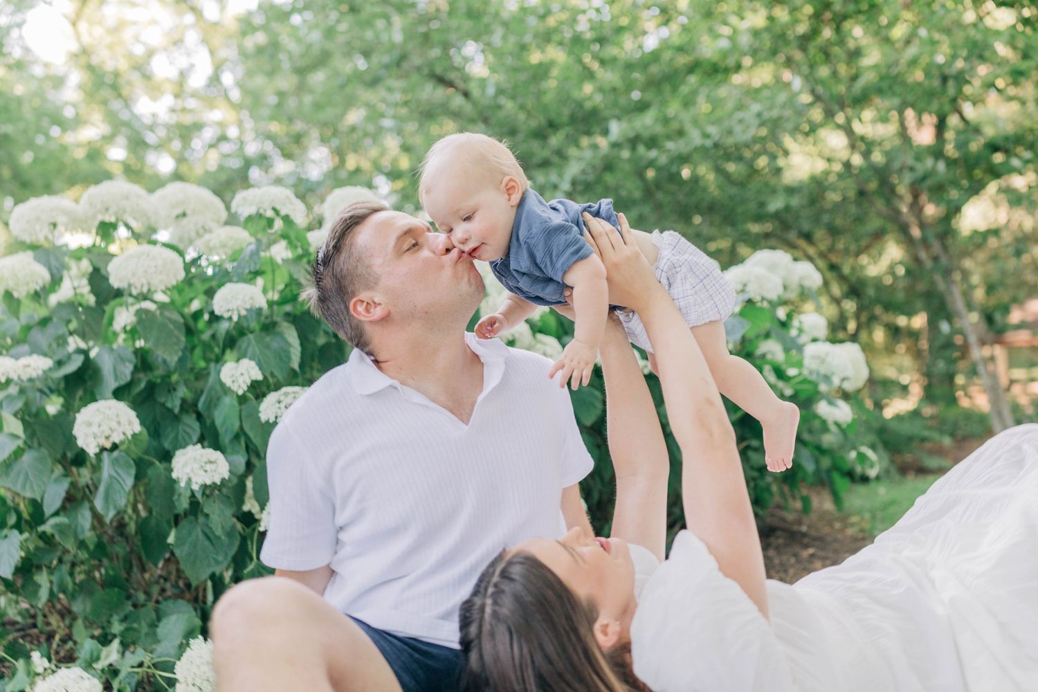 Candid family moment captured on white blanket in garden with white hydrangeas.