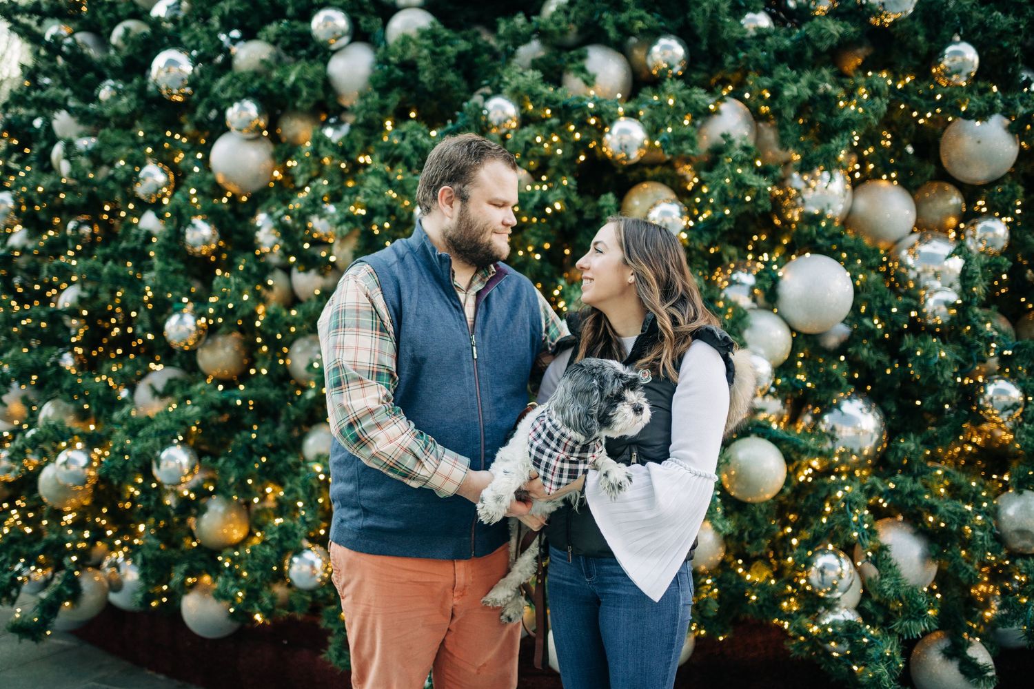 A couple shares a joyful moment in front of a Christmas tree decorated with white ornaments.