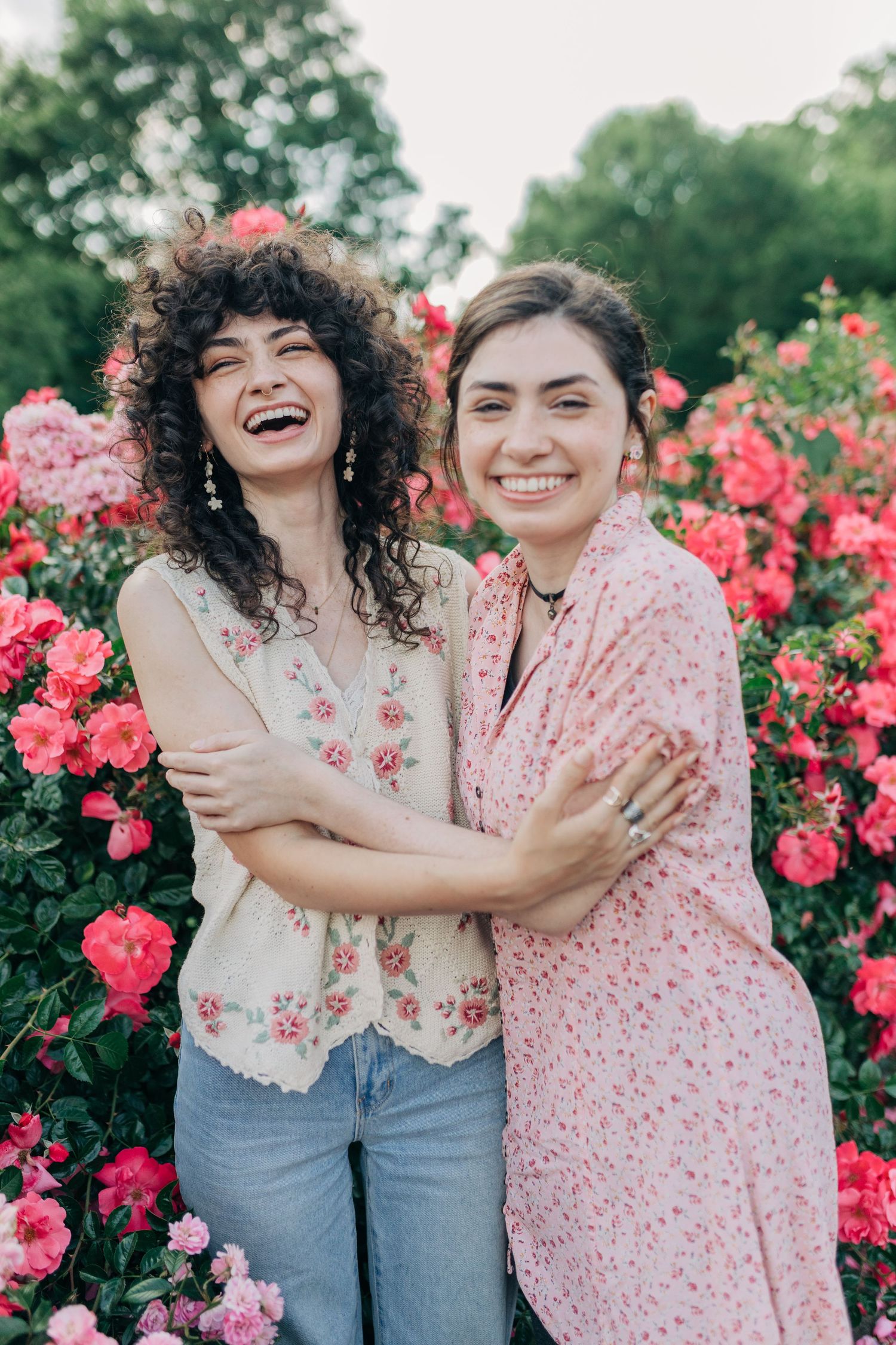 Two people wearing floral tops embrace and laugh joyfully in front of blooming pink roses in a garden.