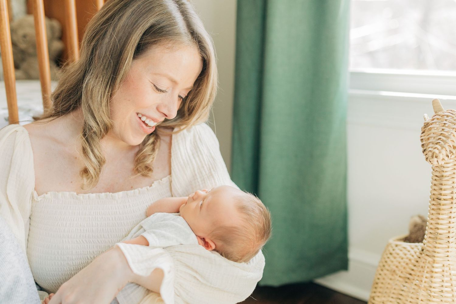 Parent in white dress cradles newborn baby in nursery with green curtains.