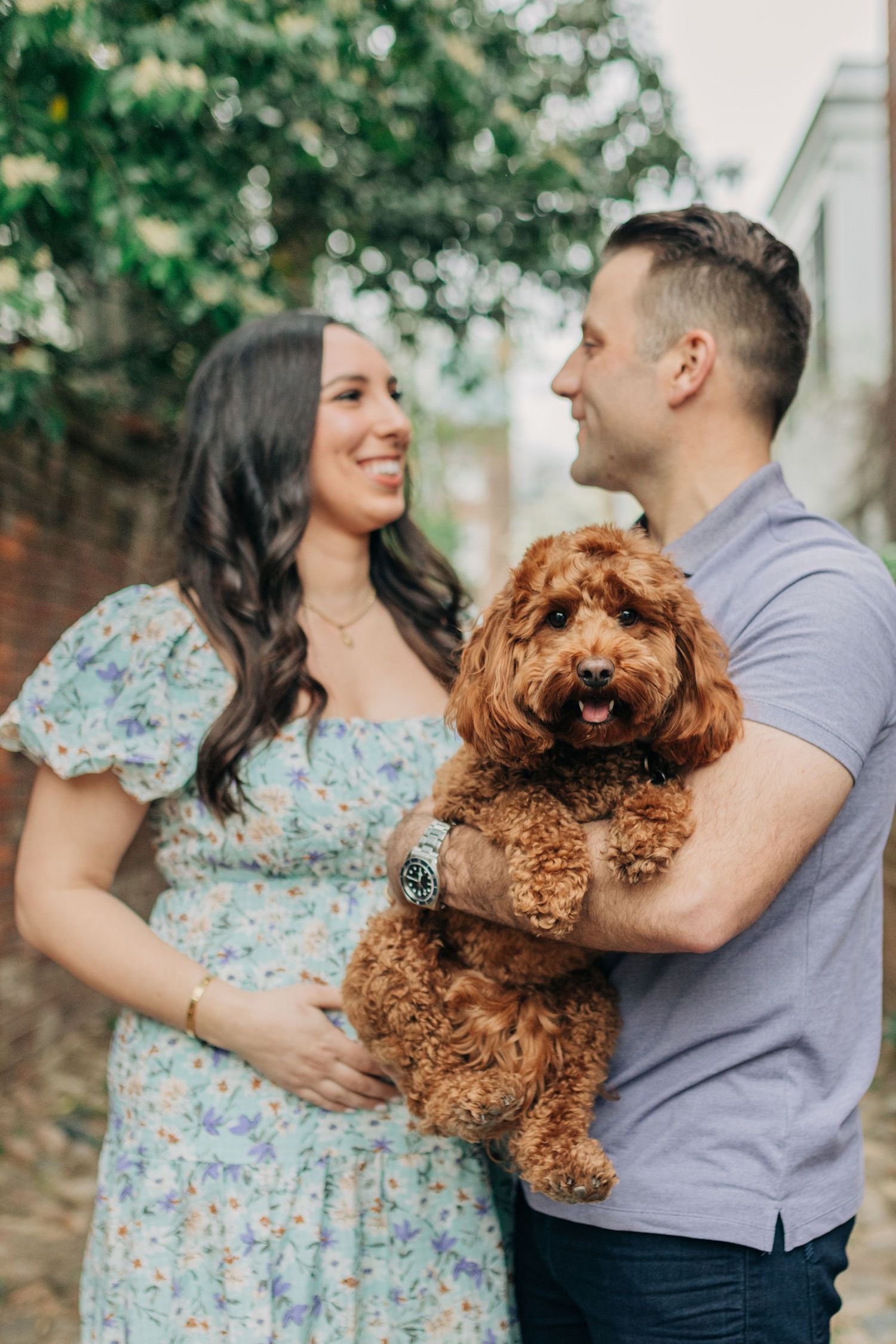 A couple shares joyful moments with their fluffy brown dog against a brick wall backdrop.