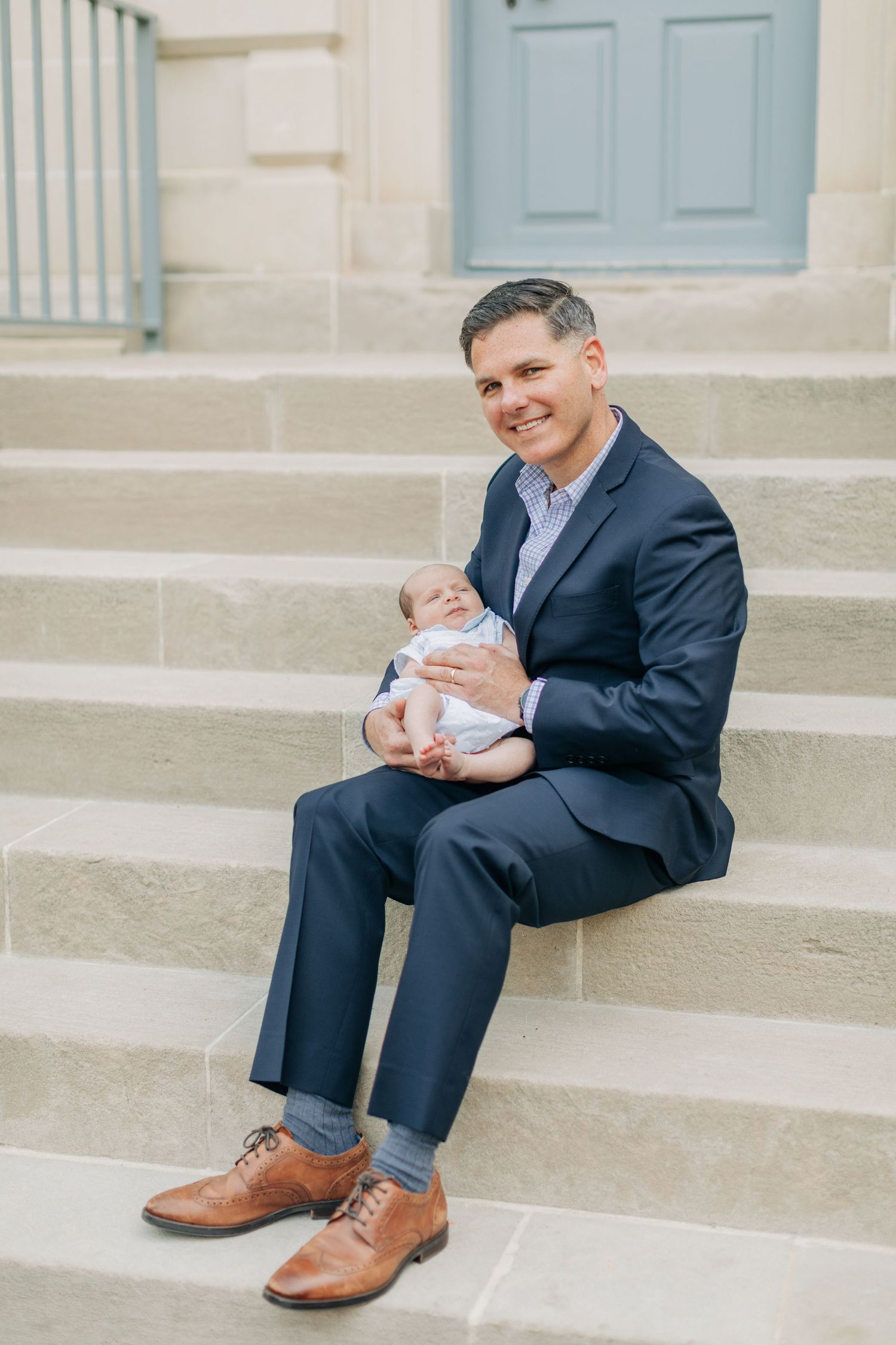 Person in navy suit and brown shoes sitting on stone steps holding an infant.