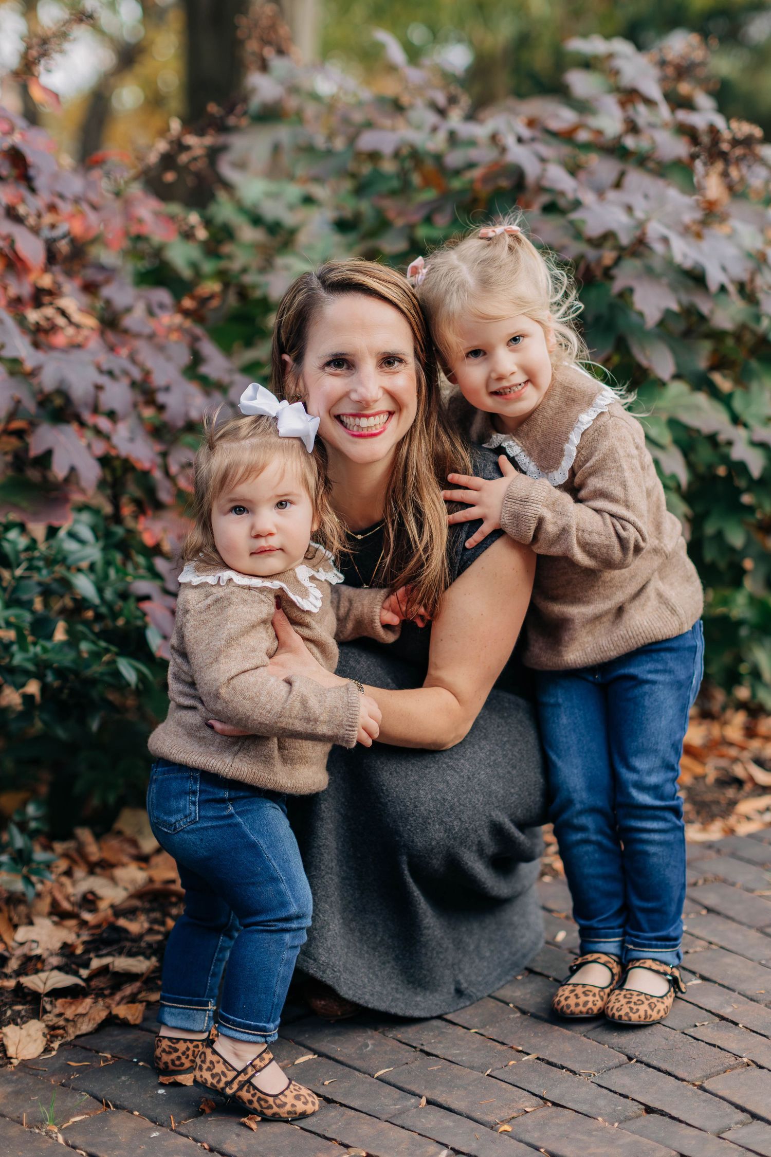 A smiling mother crouches with her two young daughters in matching tan sweaters and jeans amid autumn foliage.