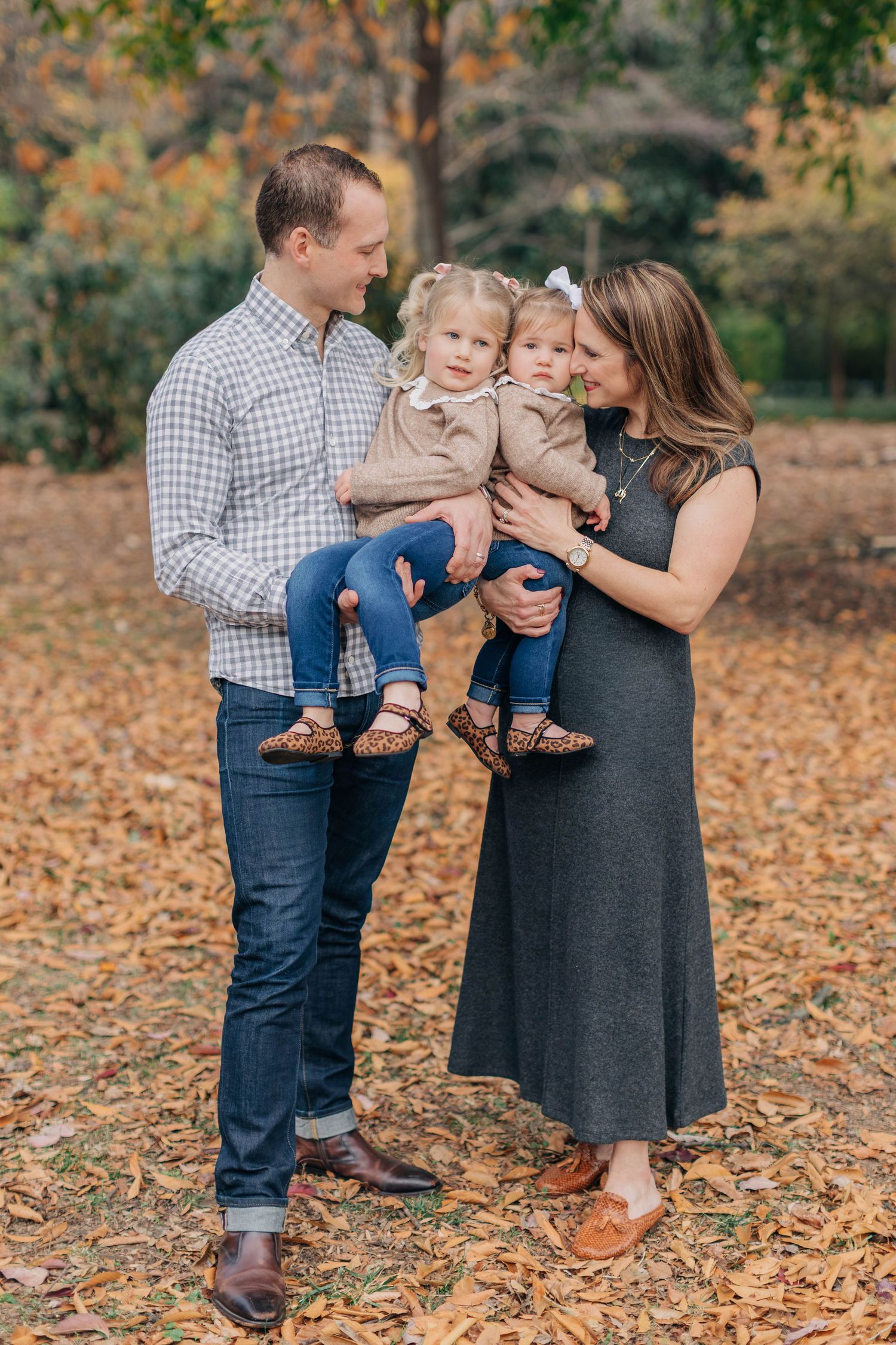 Family of four poses among autumn leaves, parents holding two toddler girls in a sunlit park.