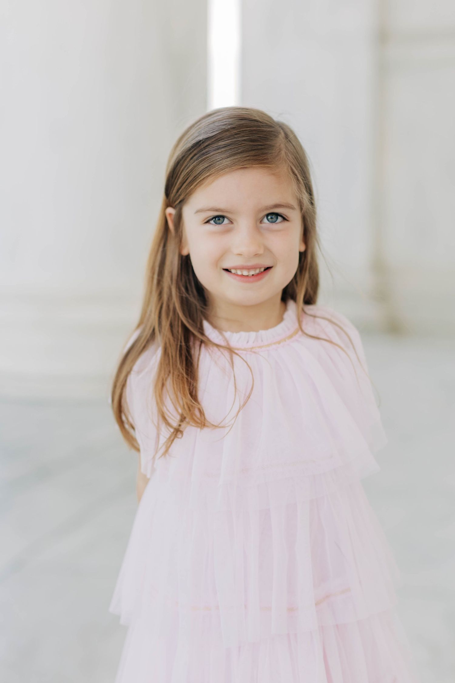 Young girl with long brown hair smiling in a pink tulle dress, standing between white columns.