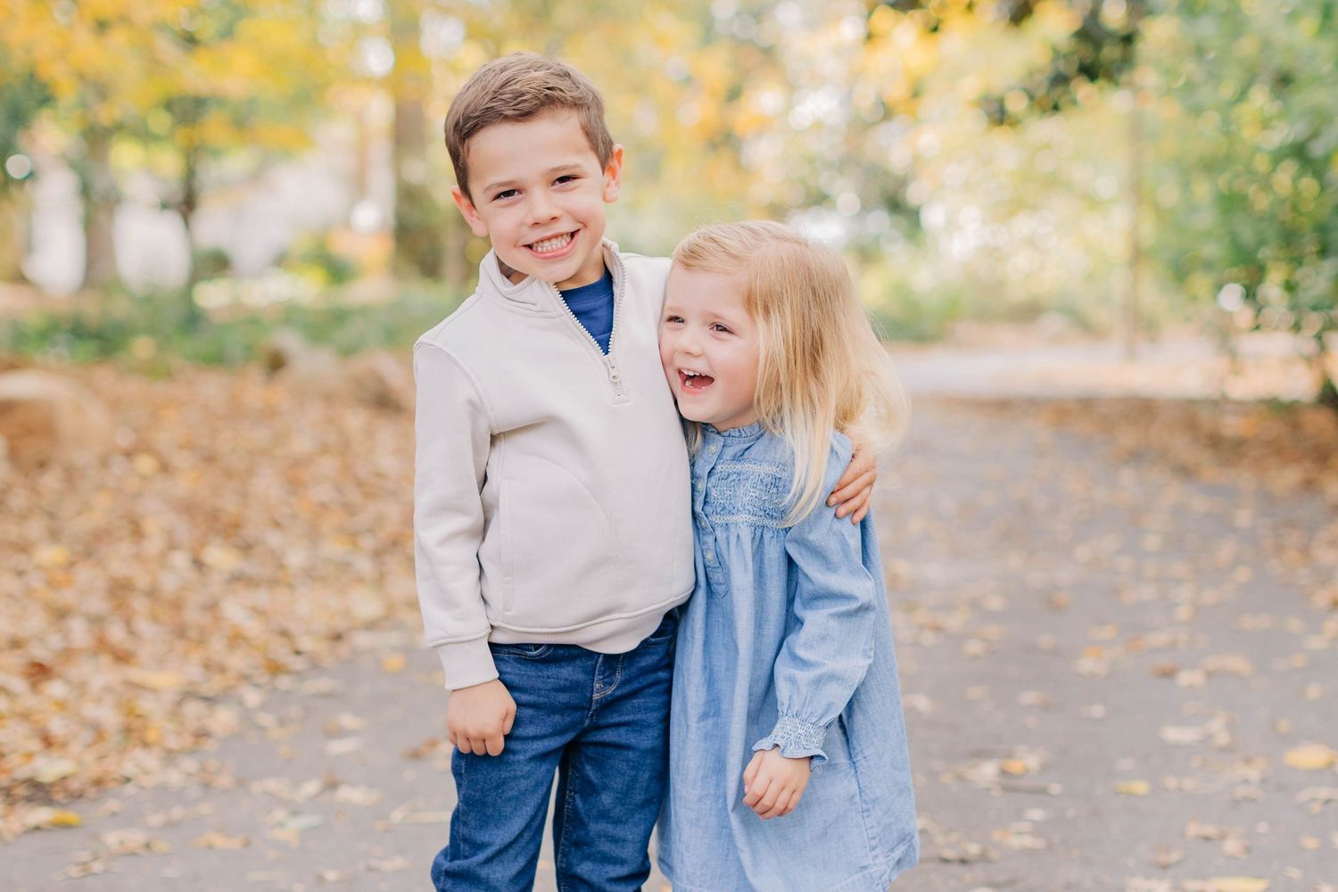 Two young siblings smiling and hugging outdoors in autumn park, boy in white jacket and girl in blue dress.
