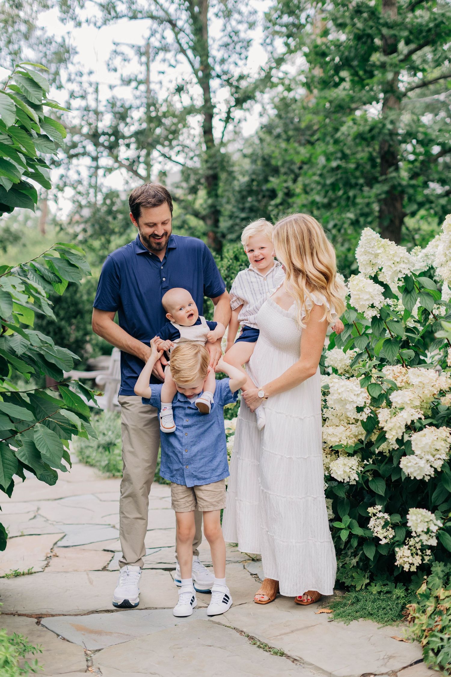 Happy family with two young children posing on a stone path surrounded by lush greenery and white hydrangeas.