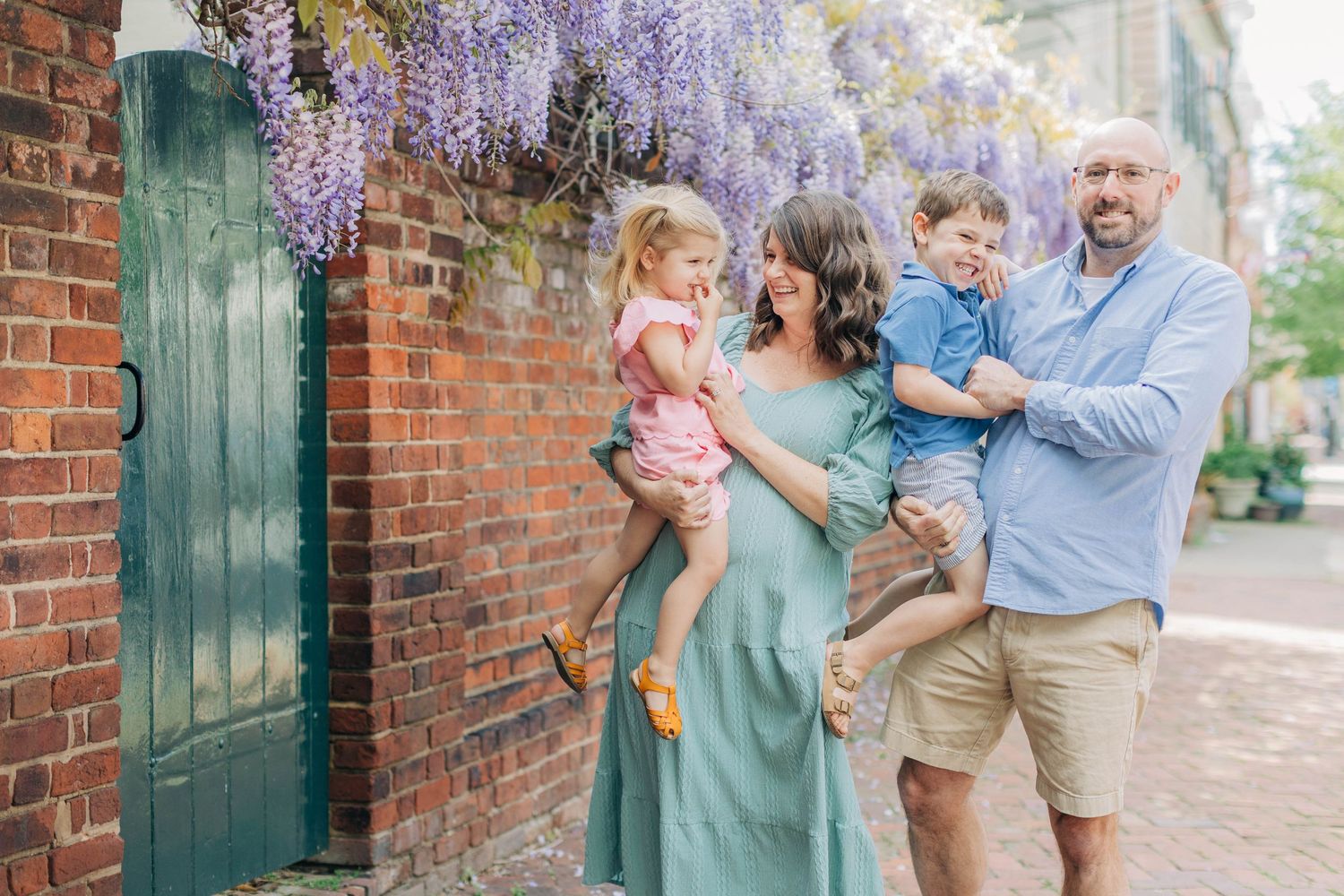 Family of four poses by brick wall with wisteria blooms, parents holding young children in charming outdoor setting.