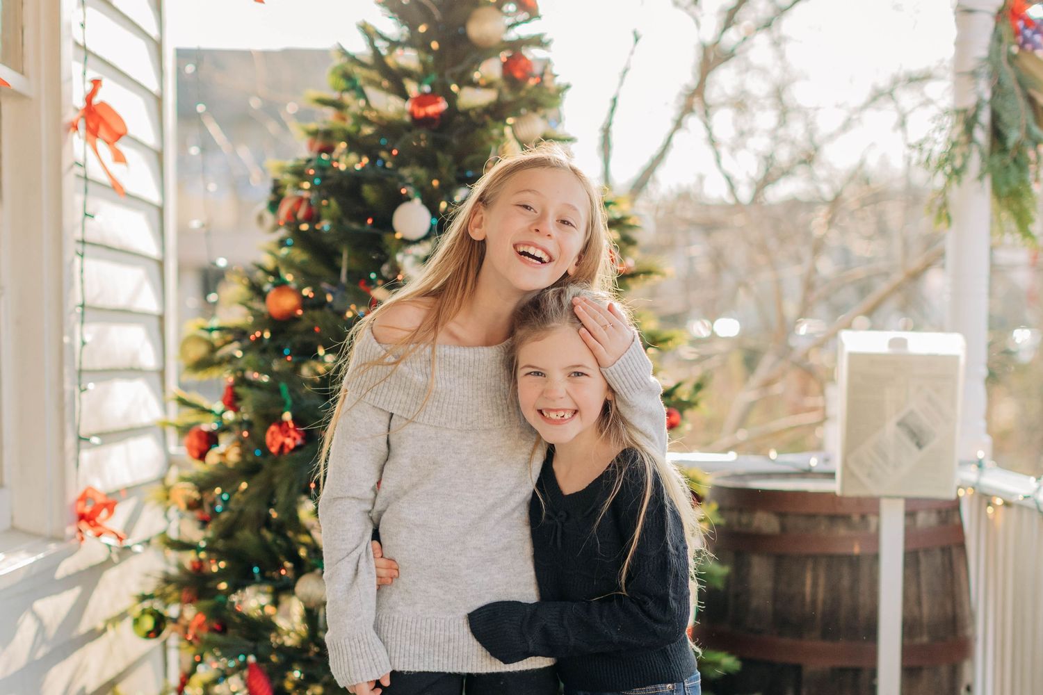 Two smiling girls hugging in front of a decorated Christmas tree on a festive porch.