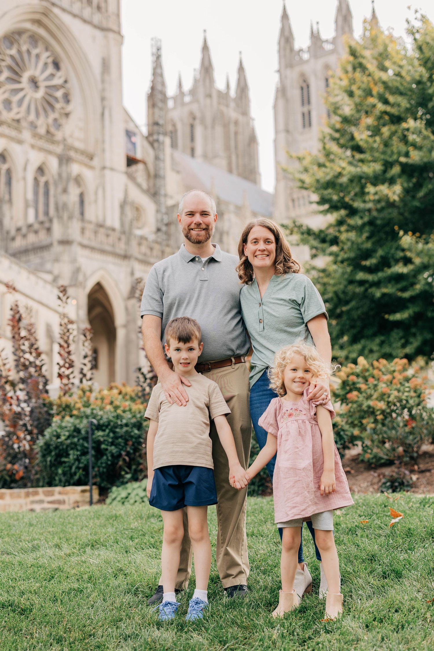 A smiling family of four stands together on a lush green lawn in front of a grand Gothic stone cathedral.