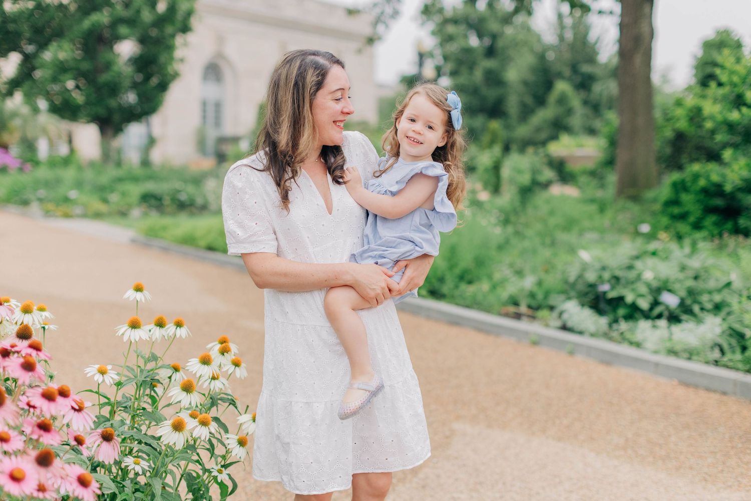 A mother in a white dress holds her young daughter in a blue outfit outdoors near colorful flowers in a garden.