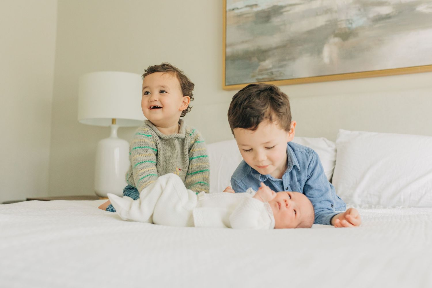 Children resting on white bedding showing cheerful expressions with wall art and lamp in background.