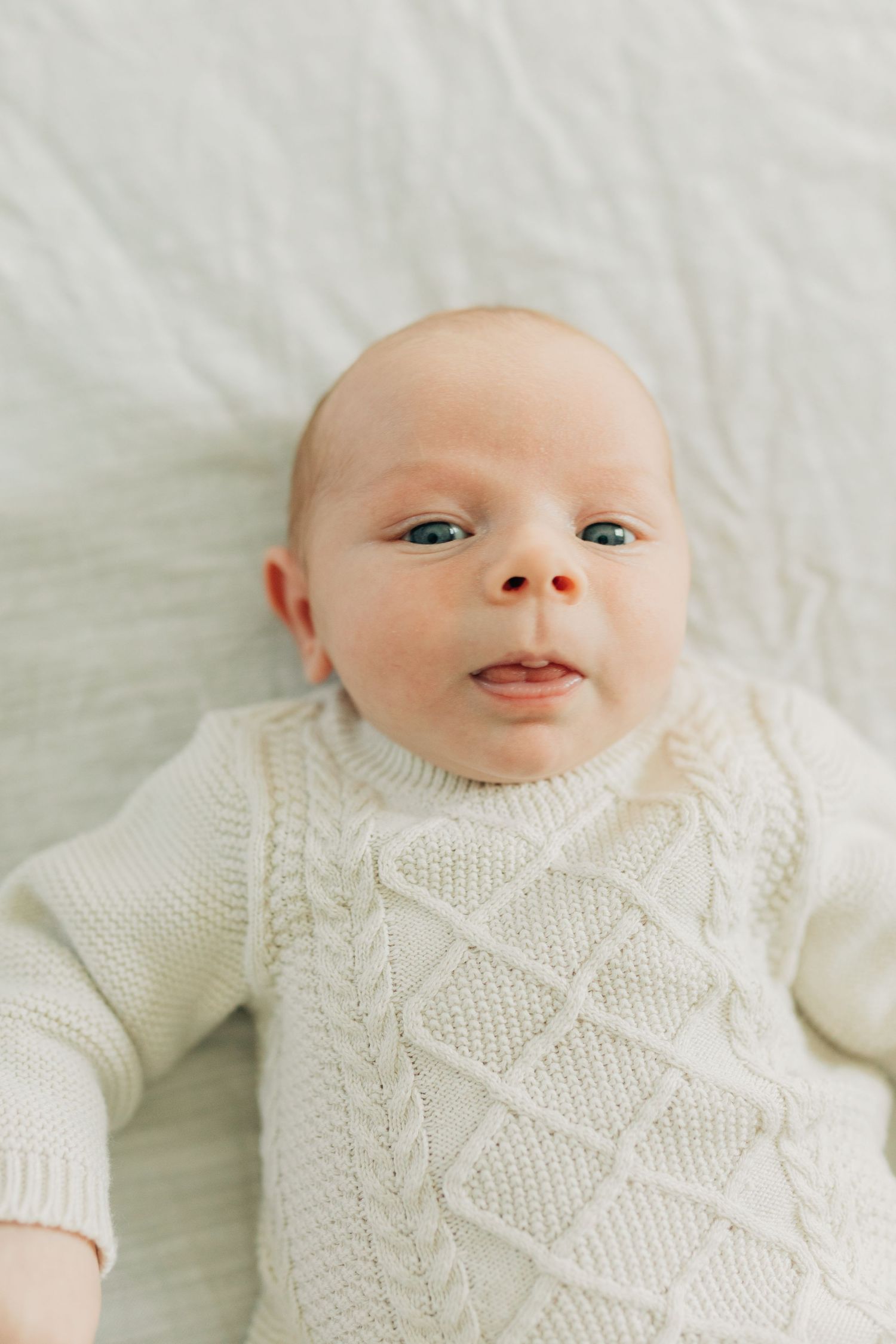 Baby in white cable knit sweater smiling on white bedding.