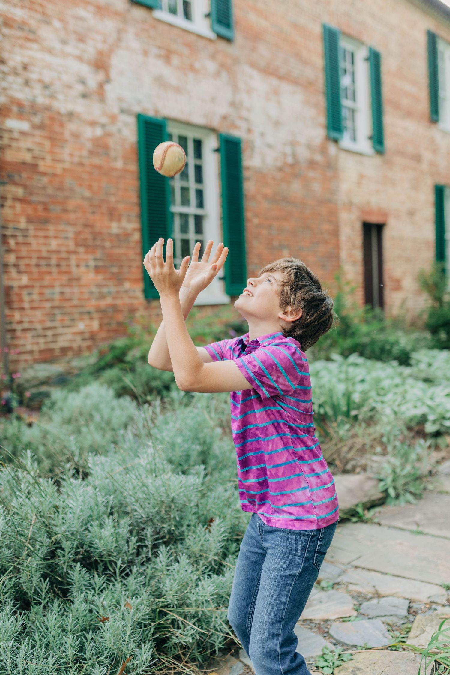 Child in striped shirt plays with ball against brick wall with green shutters and garden landscaping.