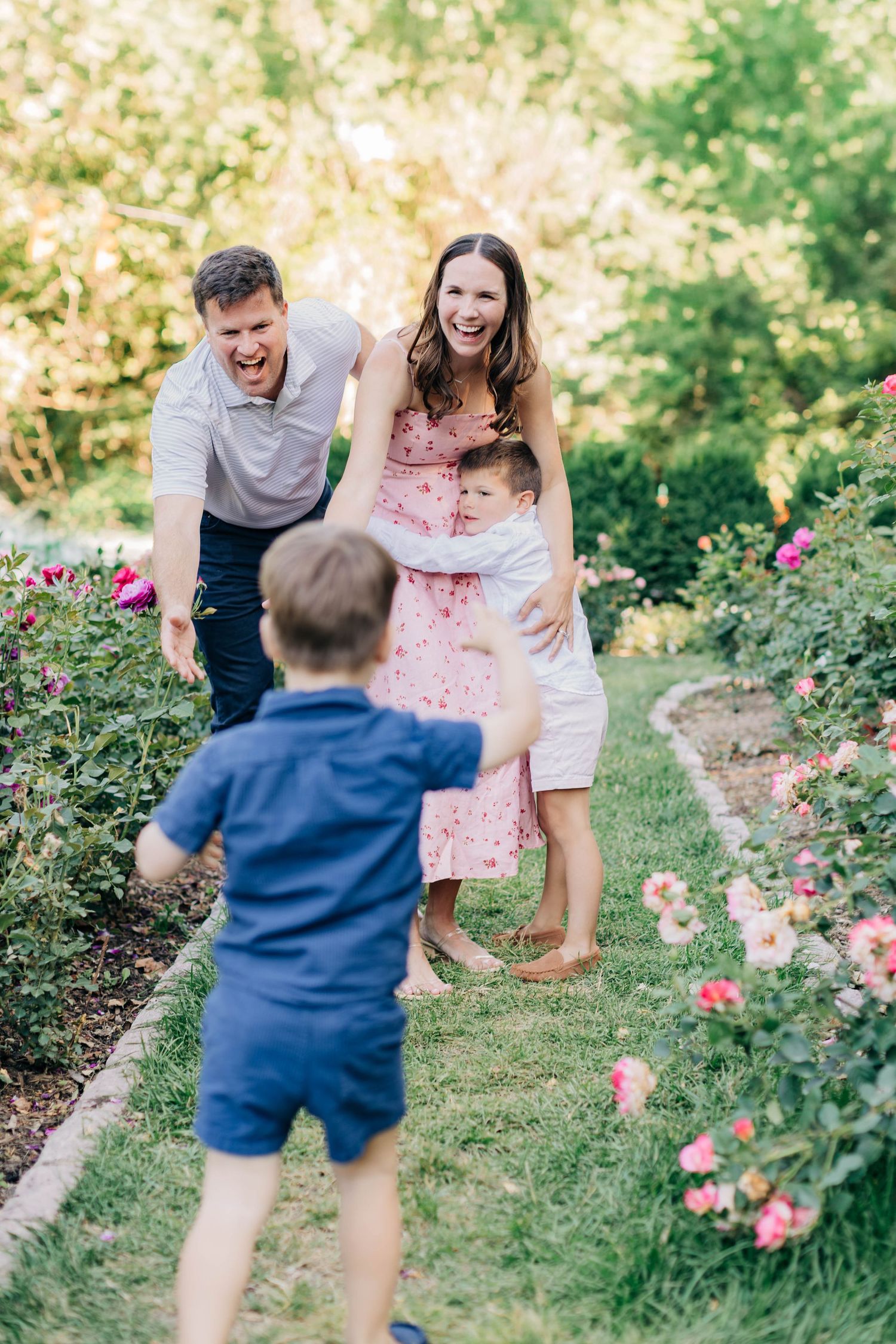 Happy family laughing as young boy runs toward parents and sibling in rose garden.